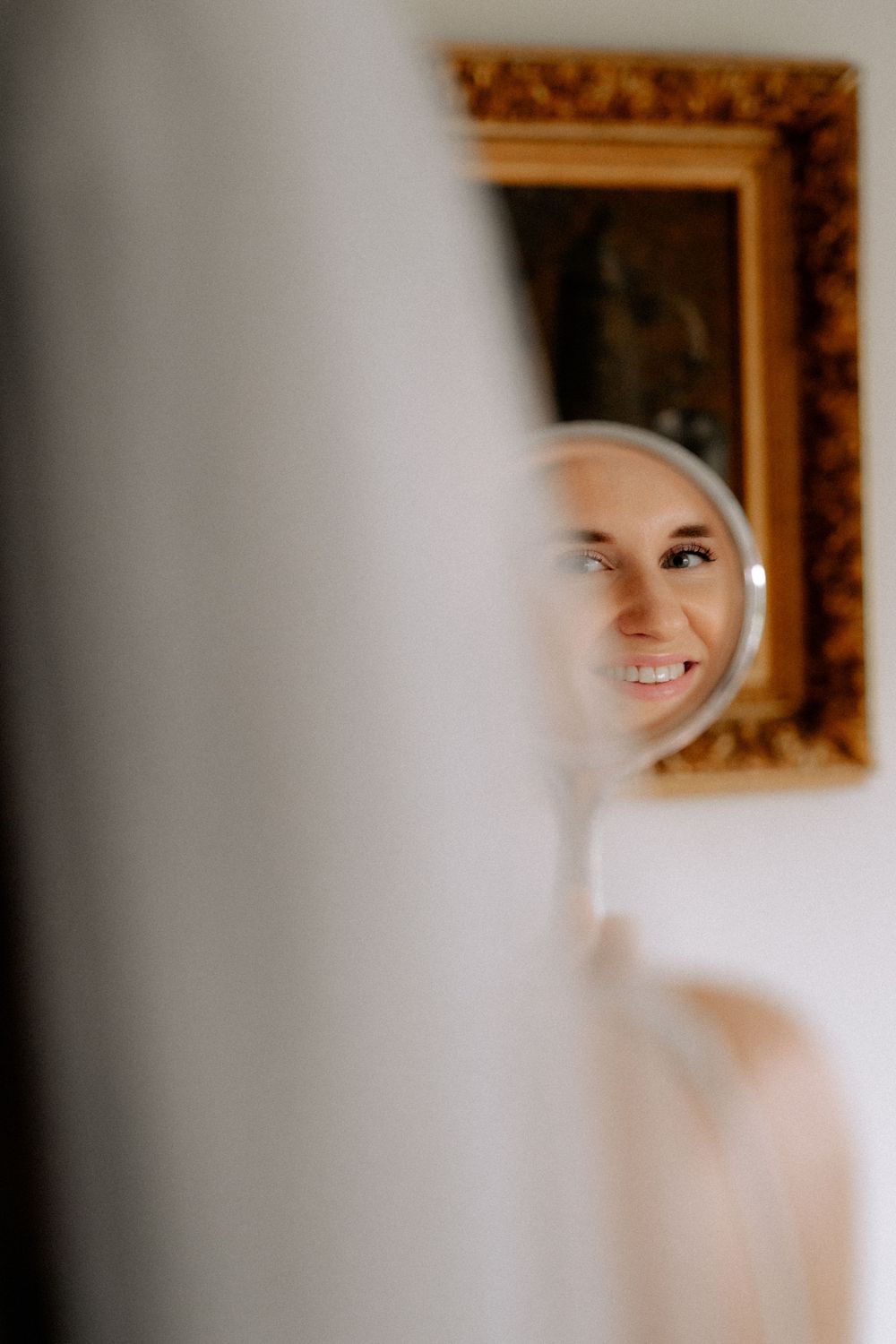 A partial reflection captures a warm smile framed by white fabric in a mirror with an ornate gold frame.