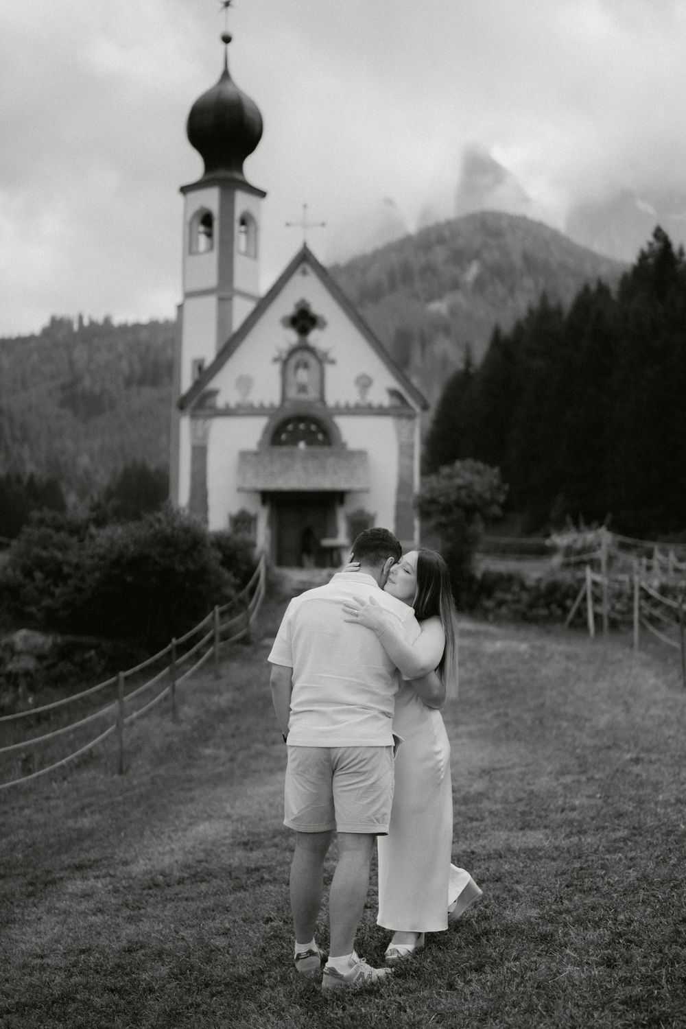 A couple walks hand in hand towards a small church nestled in the mountains, captured in black and white photography.