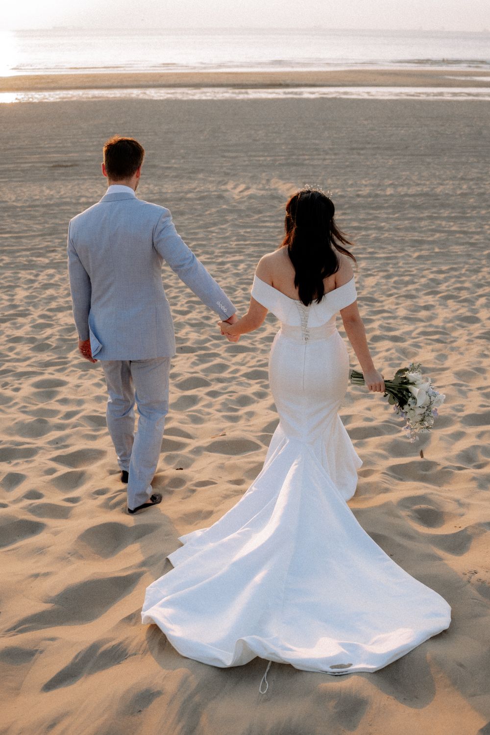Bride and groom walking hand in hand on a sandy beach at sunset, bride in white mermaid gown holding bouquet.