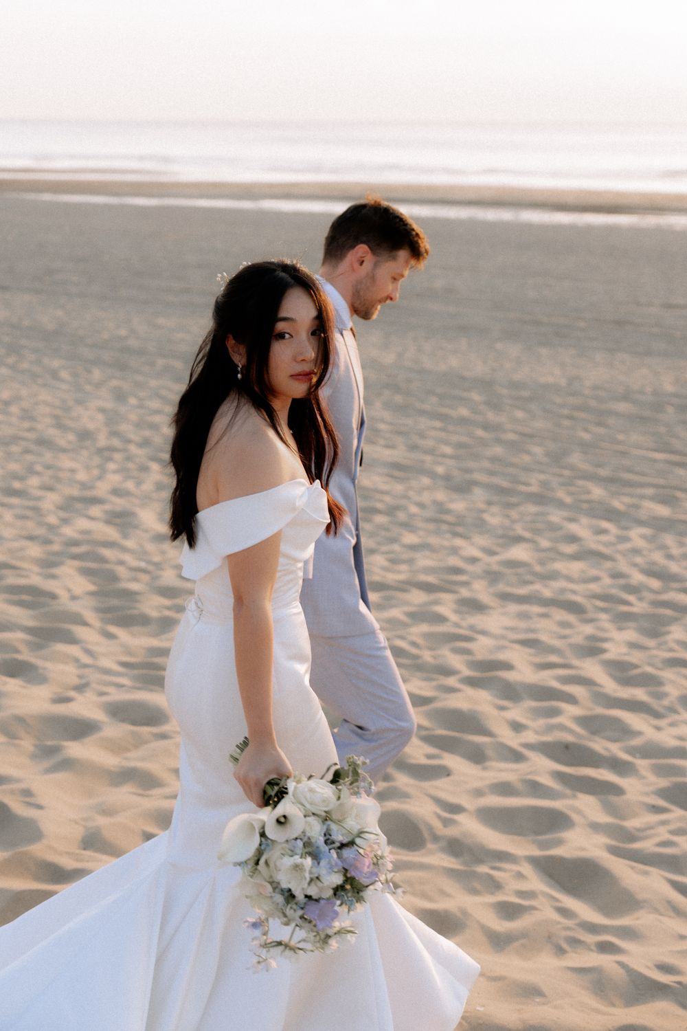 Bride in white off-shoulder gown holds bouquet on sandy beach at sunset, groom in gray suit beside her.