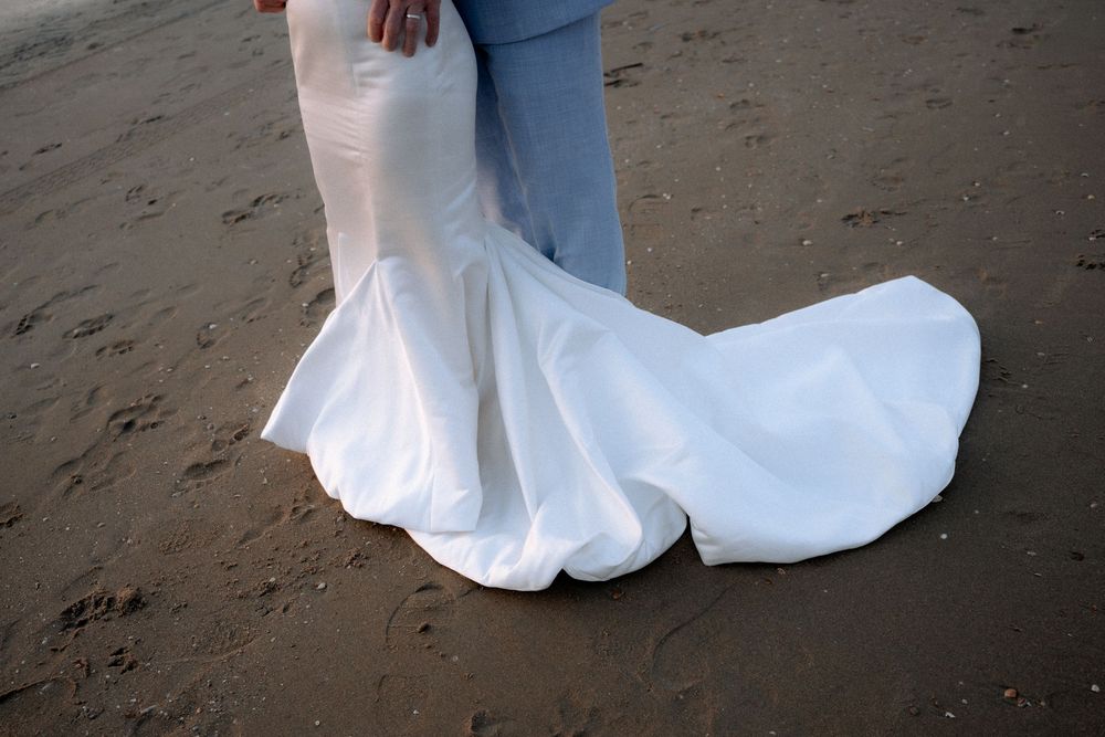 Bride in white mermaid wedding gown with flowing train standing on sandy beach beside groom in light blue pants.