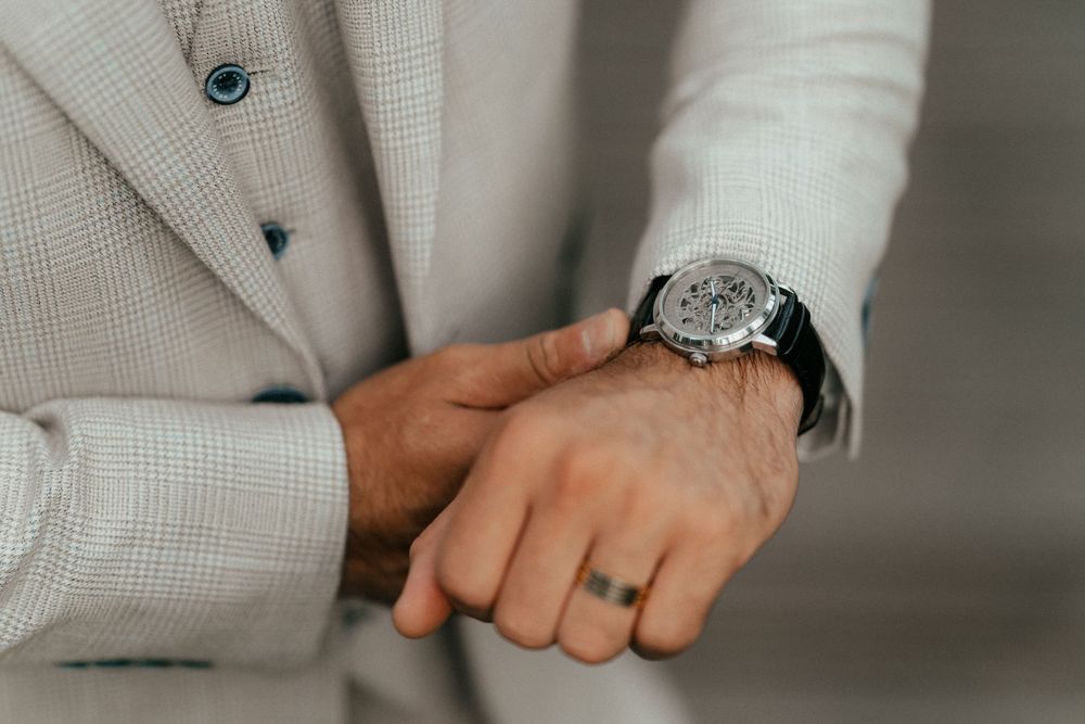 A close-up view of hands wearing a light beige suit sleeve showing an elegant wristwatch and wedding ring.