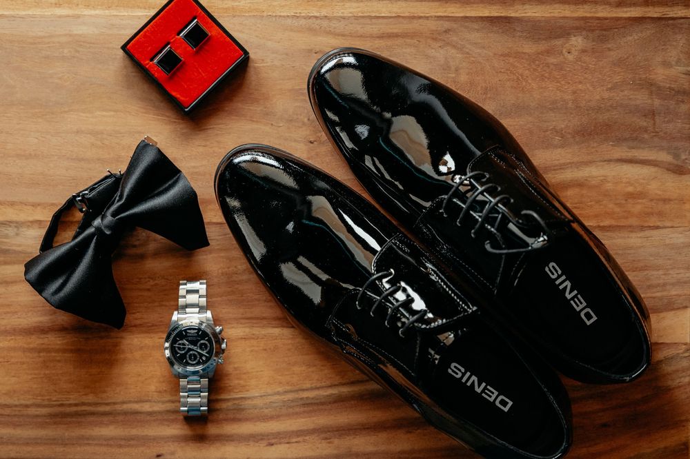 A collection of formal men's accessories including black patent dress shoes, bow tie, watch, and red ring box on wooden surface.