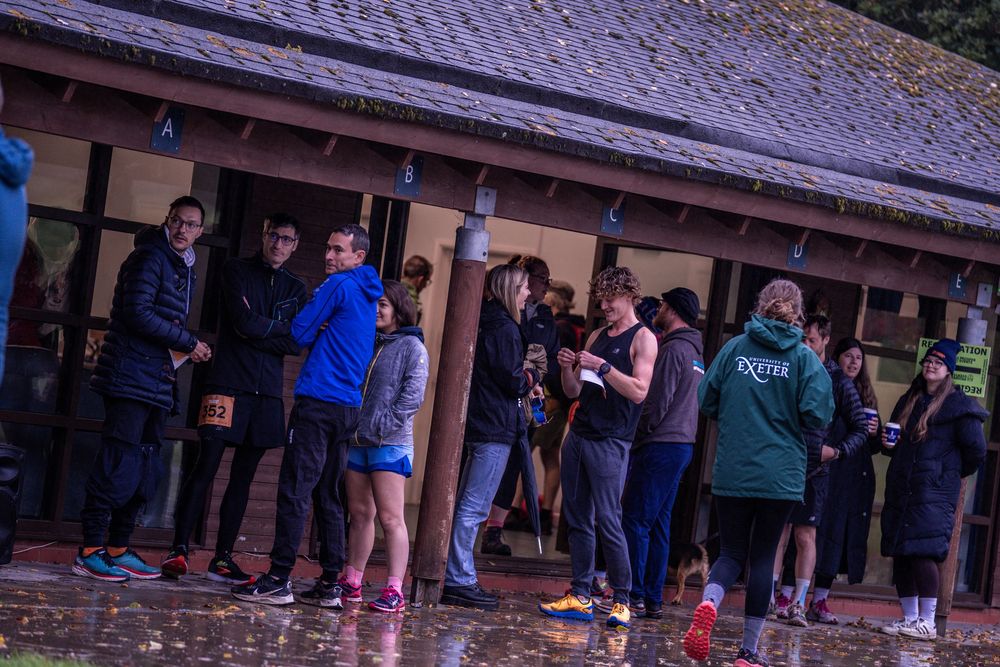Group of participants gathered outside wooden building during running event in rainy weather.