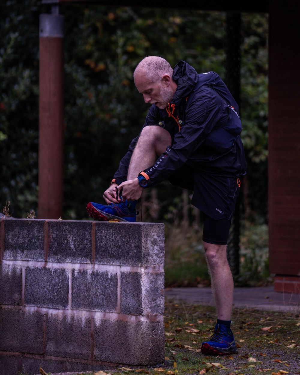 Runner adjusting shoelaces before race on concrete wall in dark clothing.