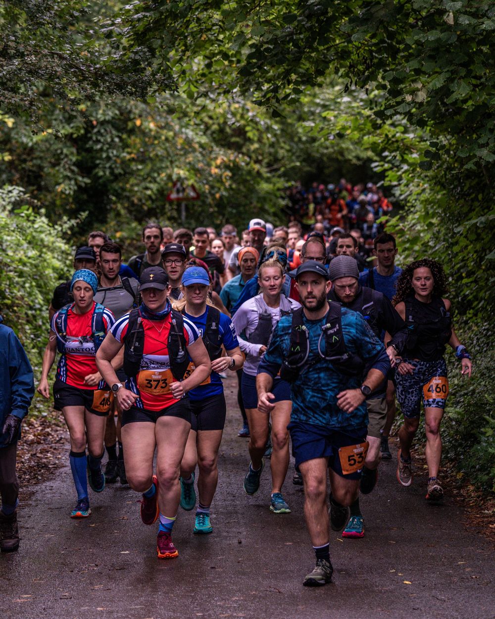 Large group of trail runners at the start of a race on a wooded path.