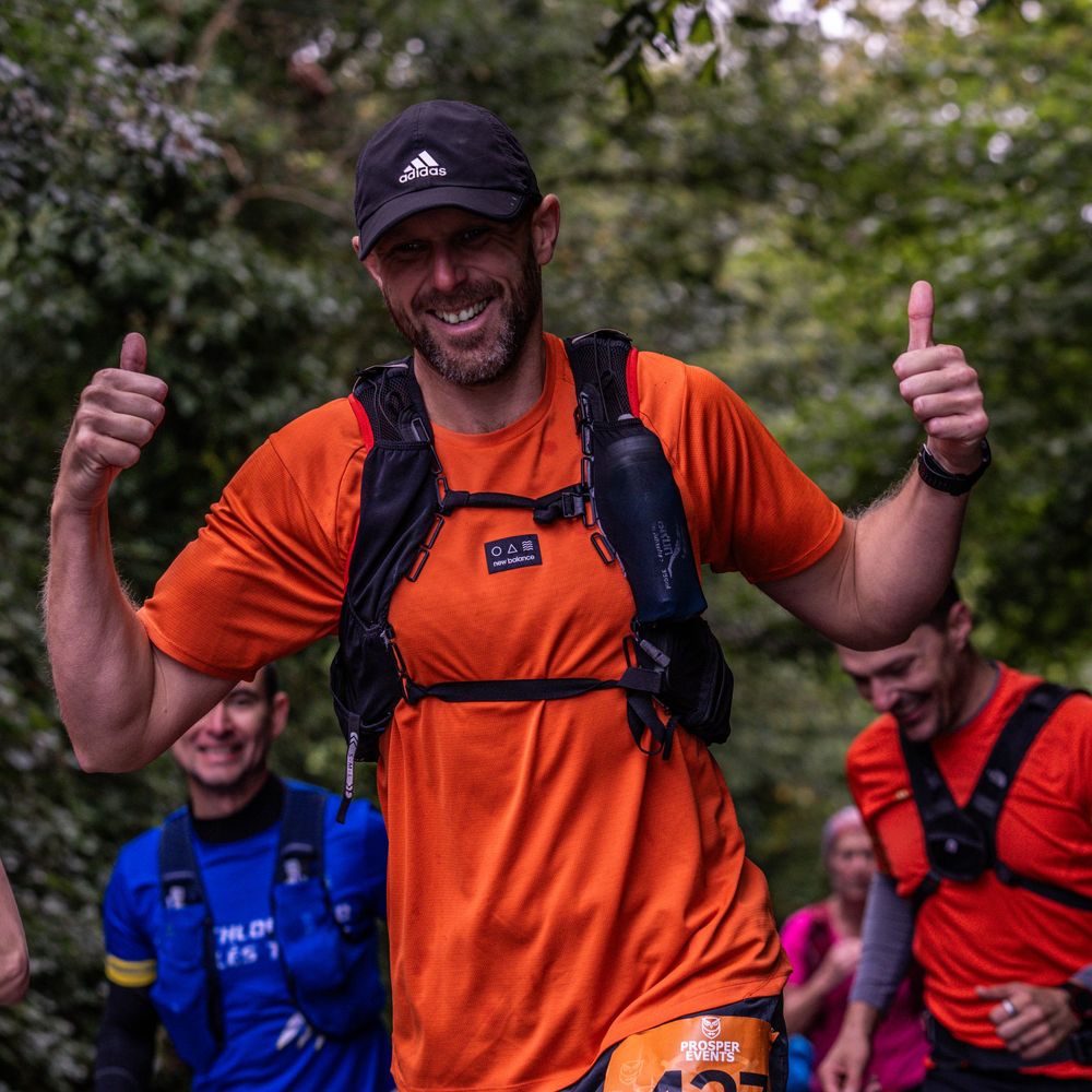 Runner in orange shirt and backpack giving thumbs up during trail race.