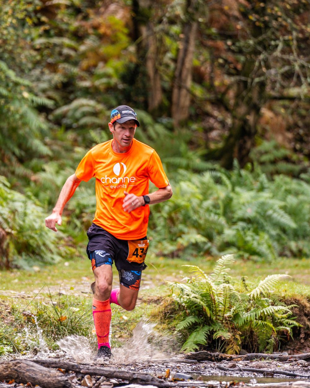 Runner in bright orange shirt splashing through stream on forest trail.