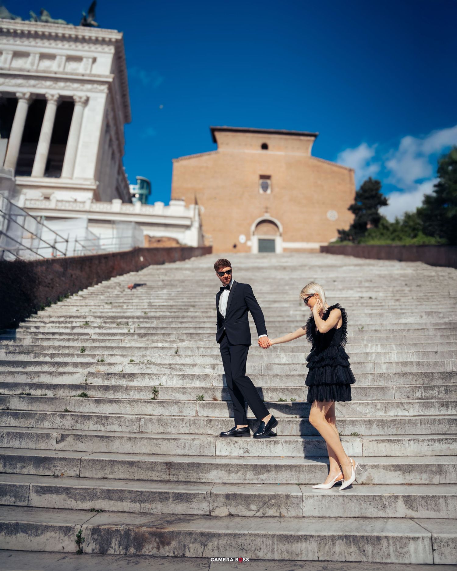A couple descends the grand stone steps of a historic building in Rome with classical architecture on a sunny day.