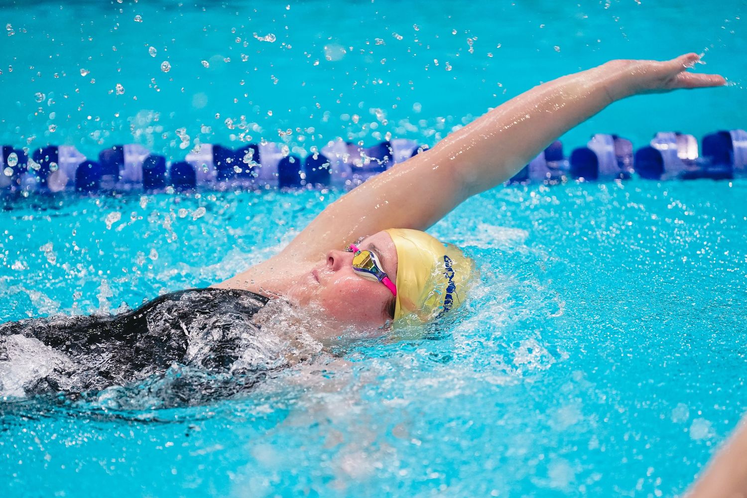 Swimmers race backstroke in a competitive pool meet with coaches and spectators watching from the sidelines.