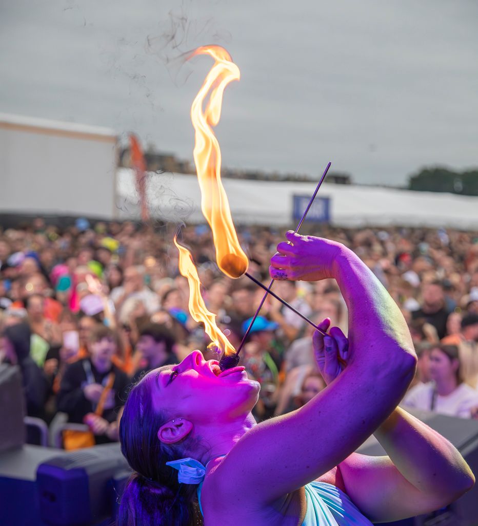 Fire performer breathes flames while entertaining a large outdoor festival crowd in vibrant purple lighting.