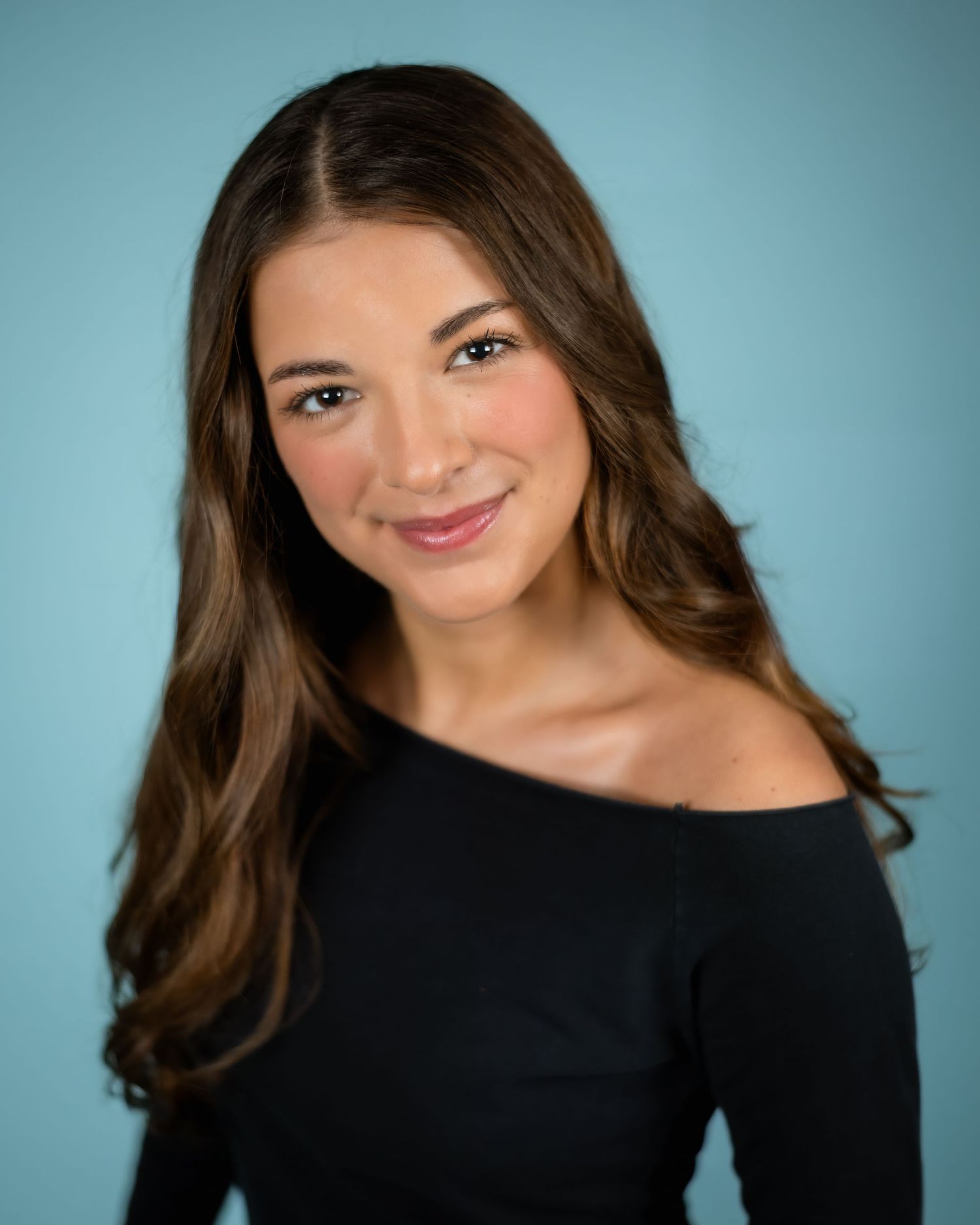 Professional headshot portrait with warm smile against blue background wearing black off-shoulder top.