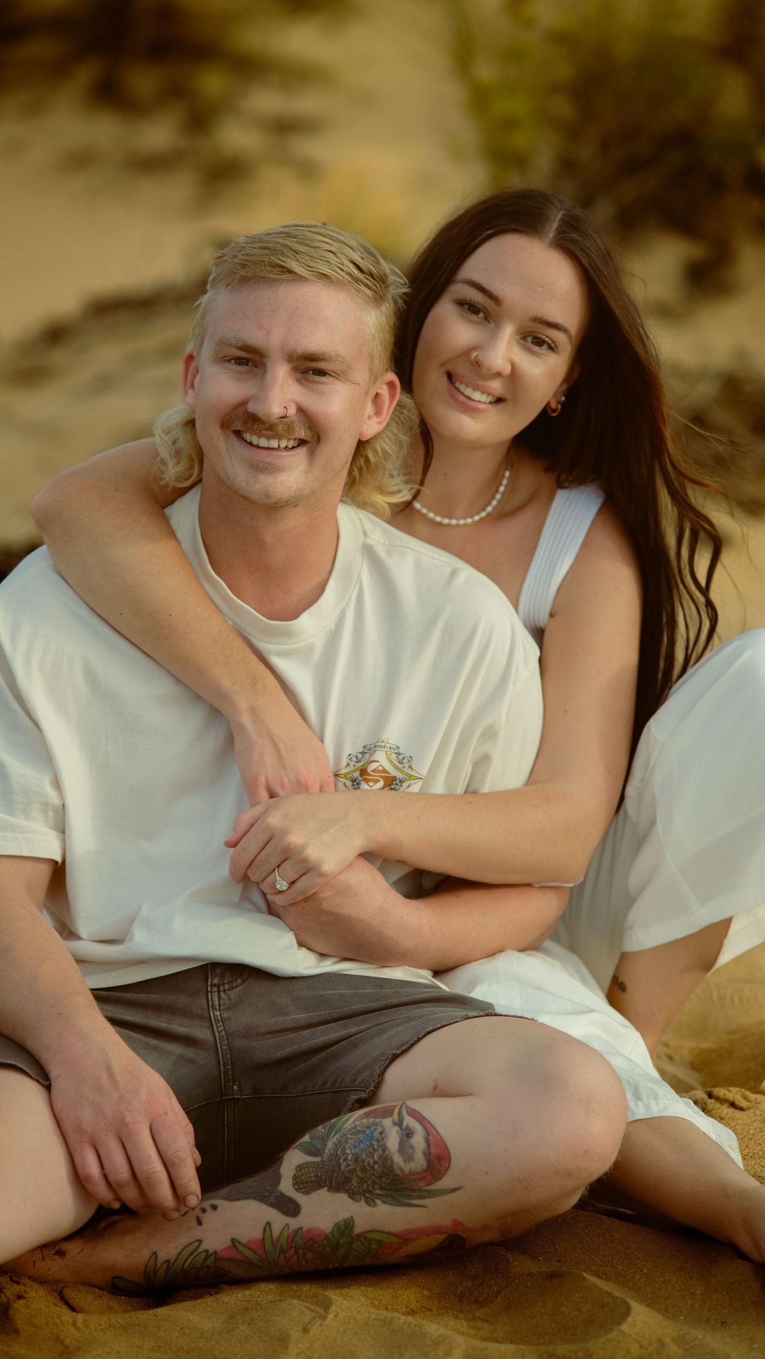 Couple relaxing on sandy beach wearing white clothing and sharing intimate moments.