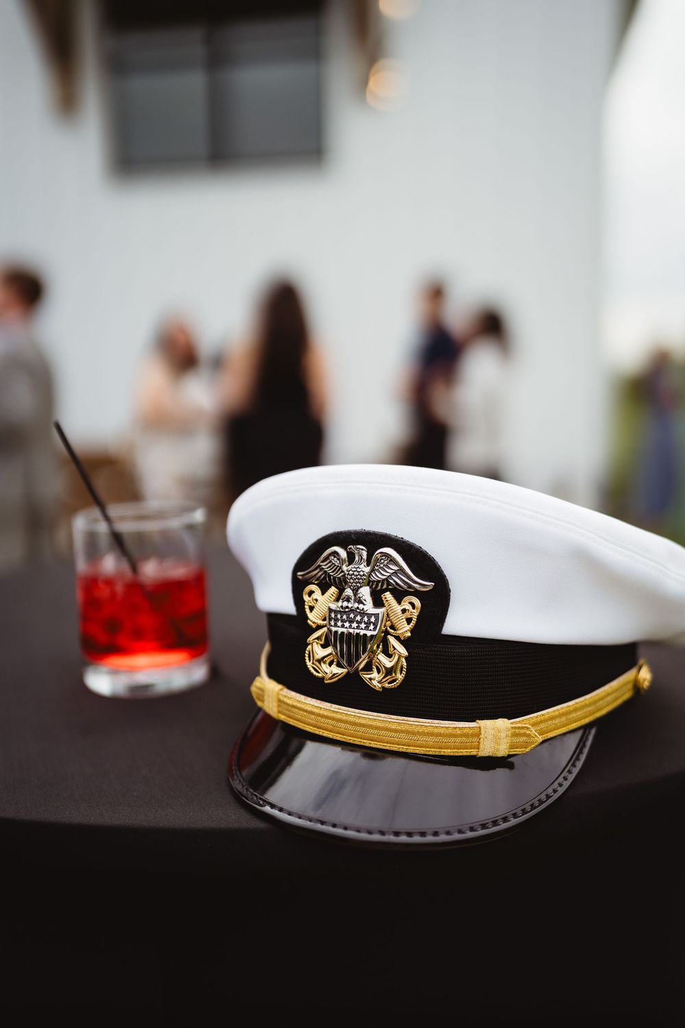 A navy captain's hat with gold trim and emblem sits on a dark table next to a red cocktail drink.