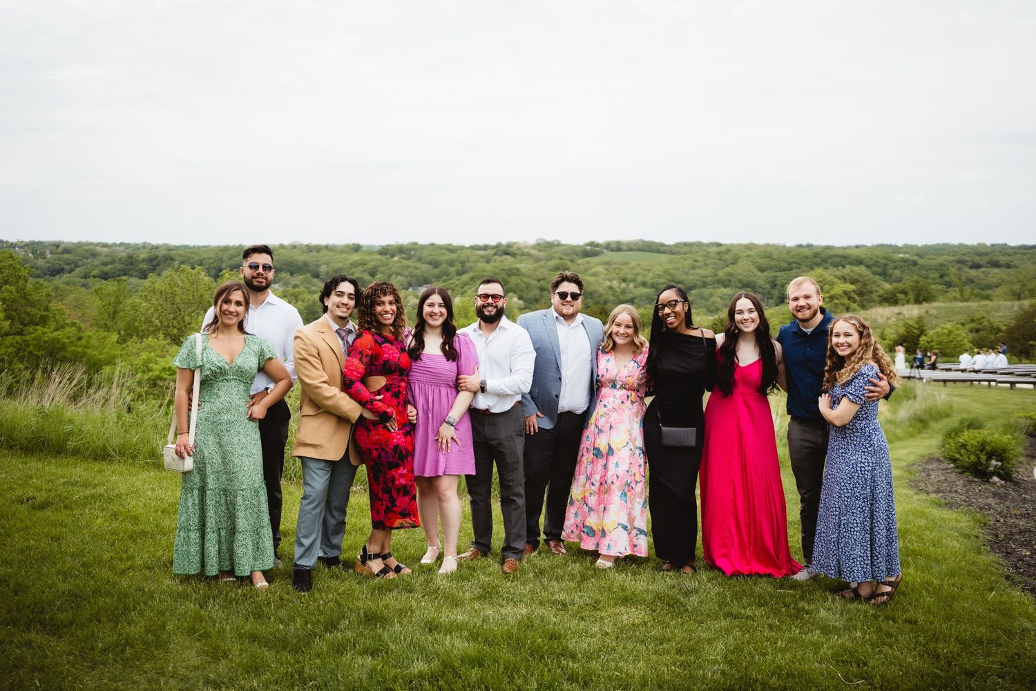 A group of wedding guests in colorful attire pose together for a photo on a grassy hill.
