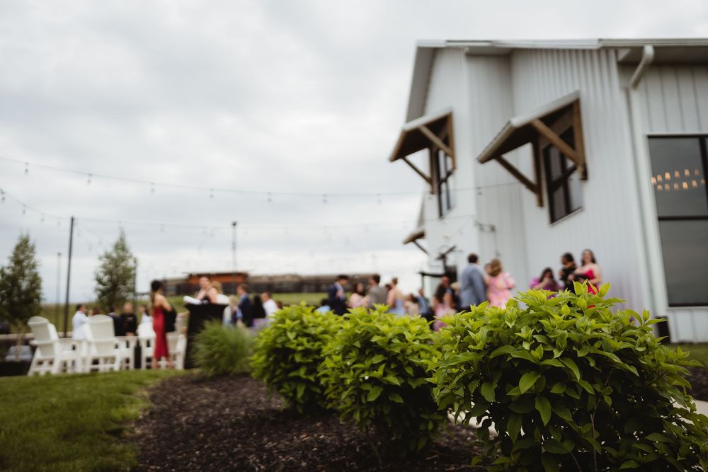 Guests mingle outside a rustic barn venue with white chairs and lush greenery in the foreground during an outdoor gathering.