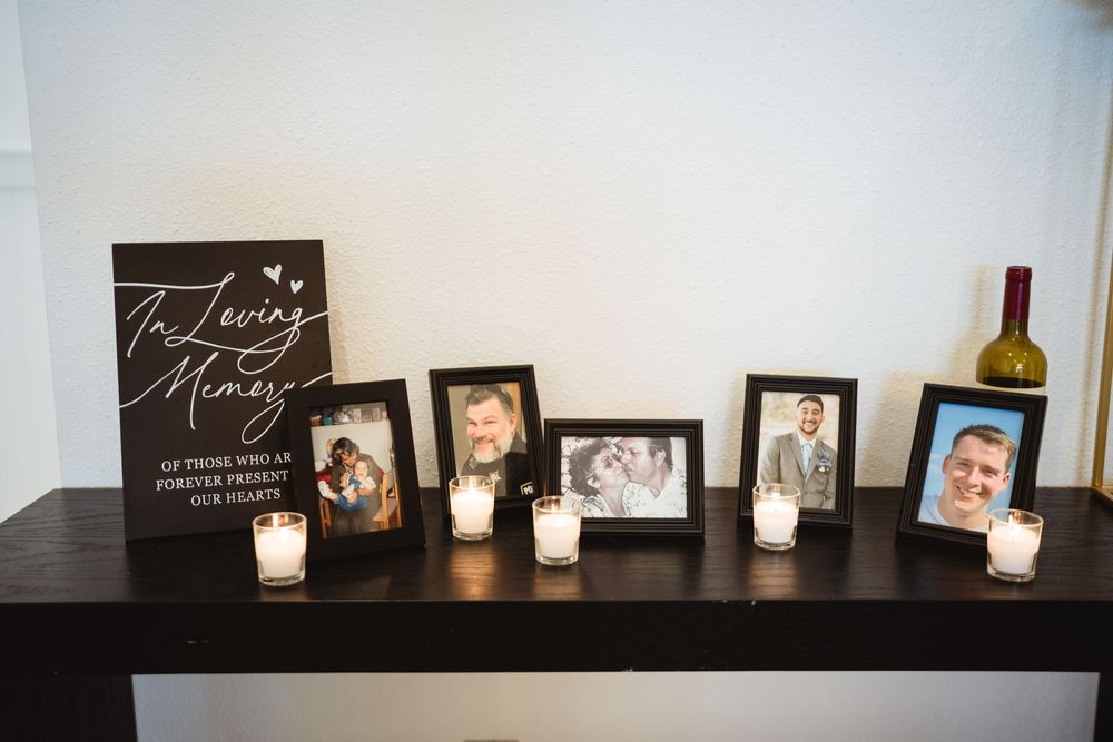 A row of lit candles sits on a dark shelf alongside framed photographs and decor items in a dimly lit room.