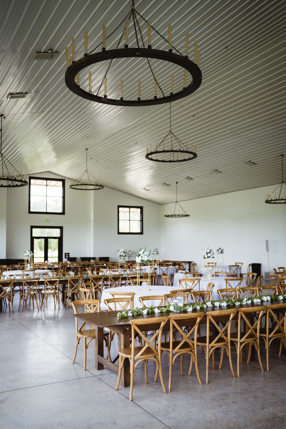 Large rustic wedding reception venue with wooden cross-back chairs and round chandeliers.