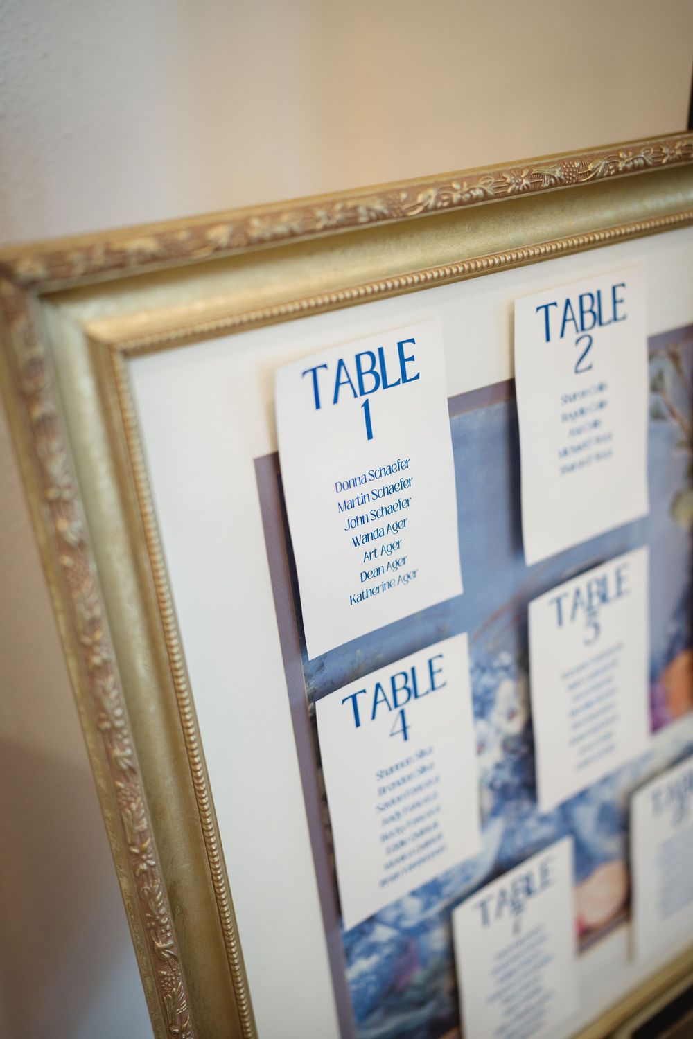 An ornate gold frame displays wedding reception table seating assignments in blue text against a white background.