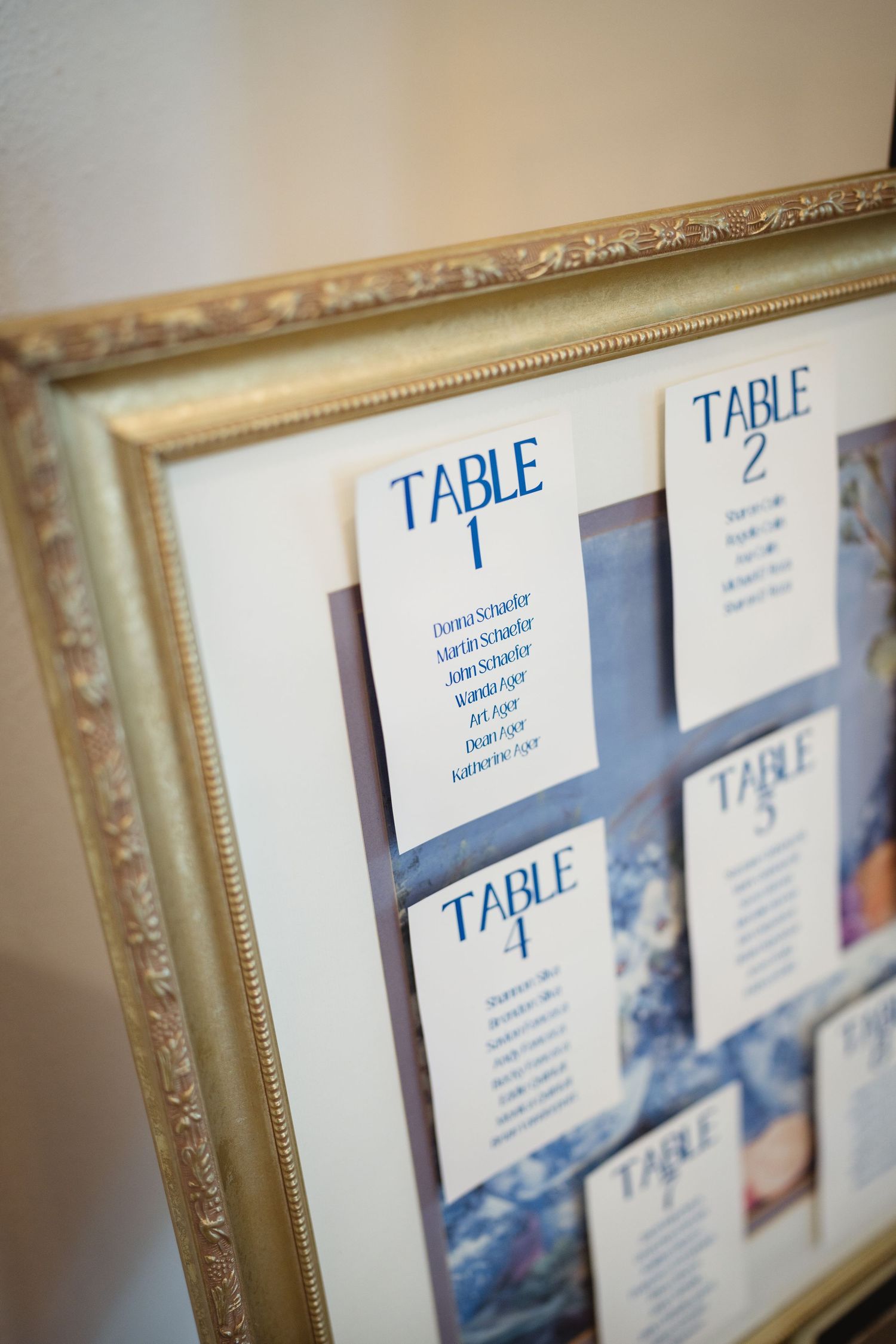 An ornate gold frame displays wedding reception table seating assignments in blue text against a white background.