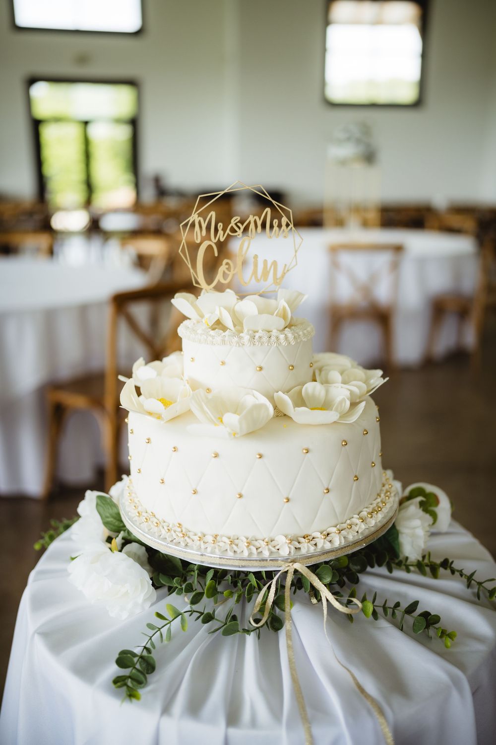 White two-tier wedding cake decorated with roses and gold accents on white linen with eucalyptus greenery.