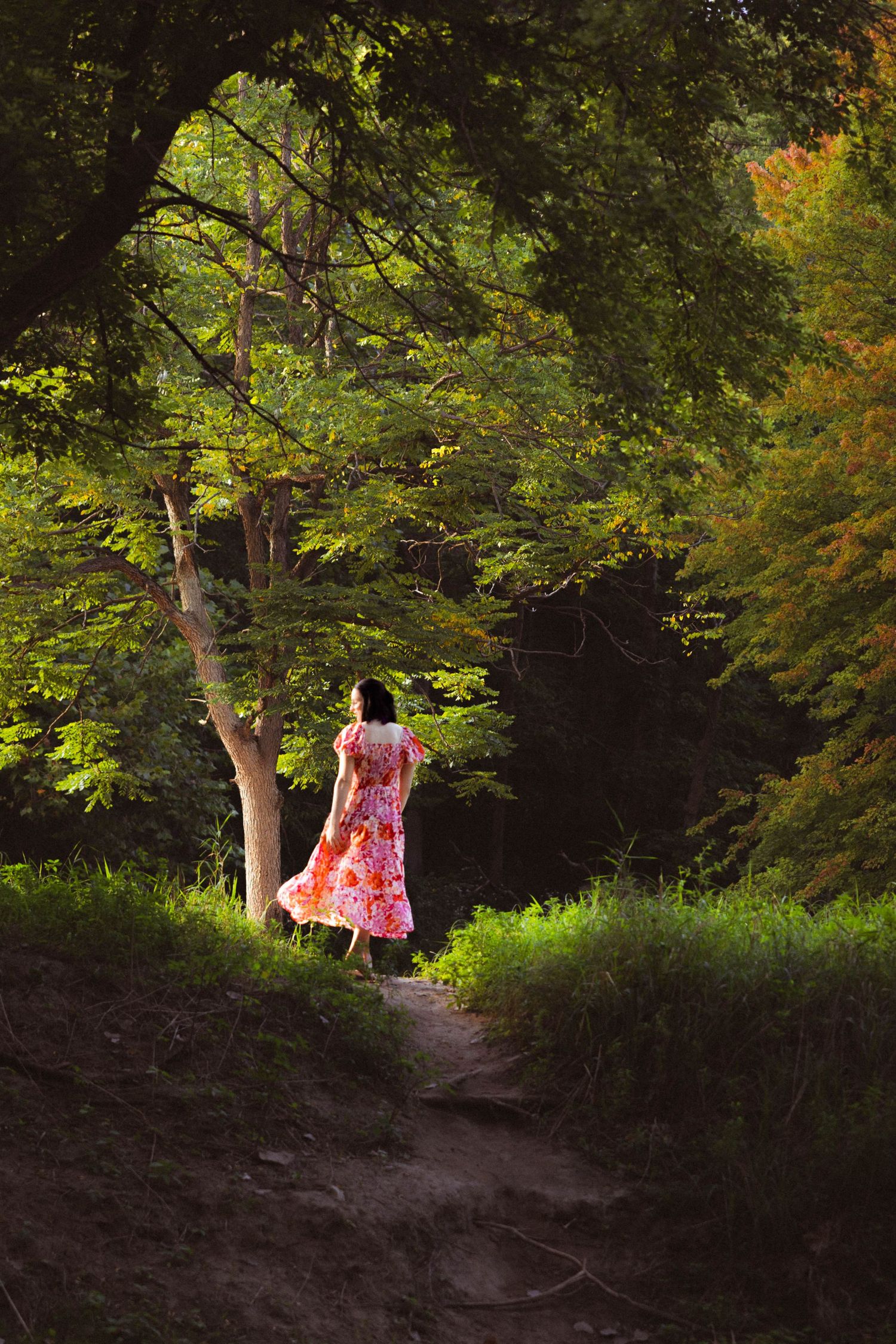 A figure in a flowing pink dress walks along a shaded forest path surrounded by lush green trees and dappled sunlight.