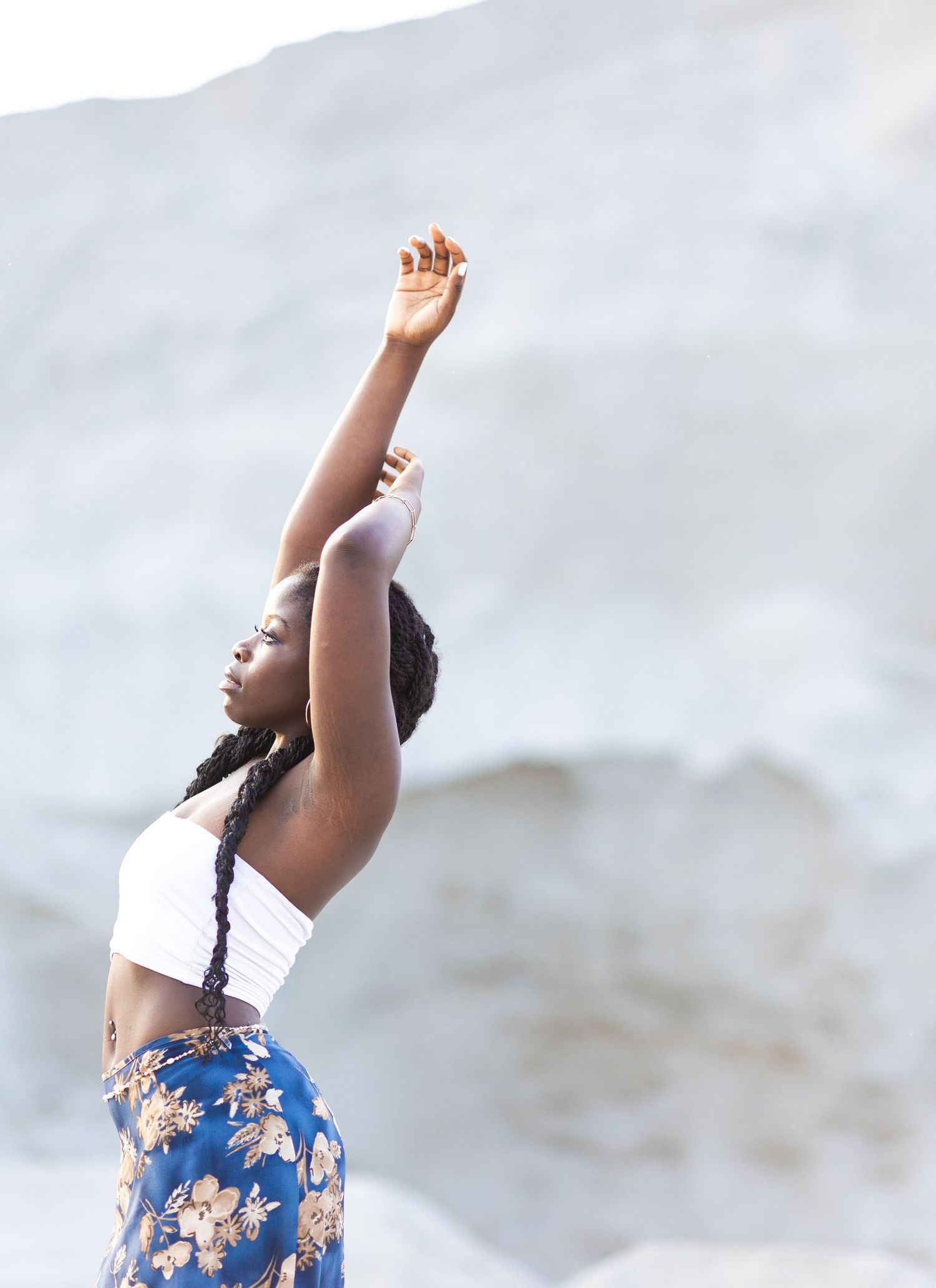 Person in white top and floral blue skirt stretches upward with raised arms against a light background.