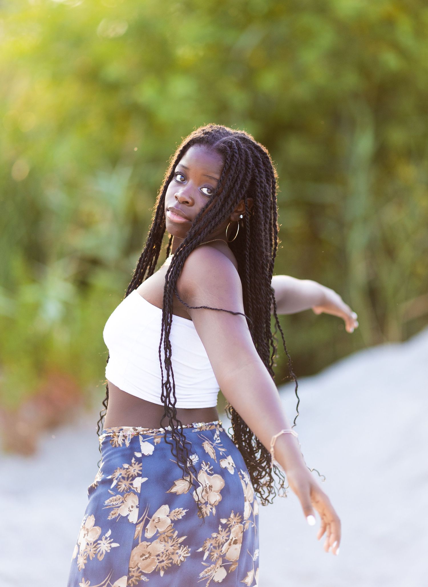 A graceful dancer in a white crop top and floral skirt moves expressively against a soft, blurred natural background.