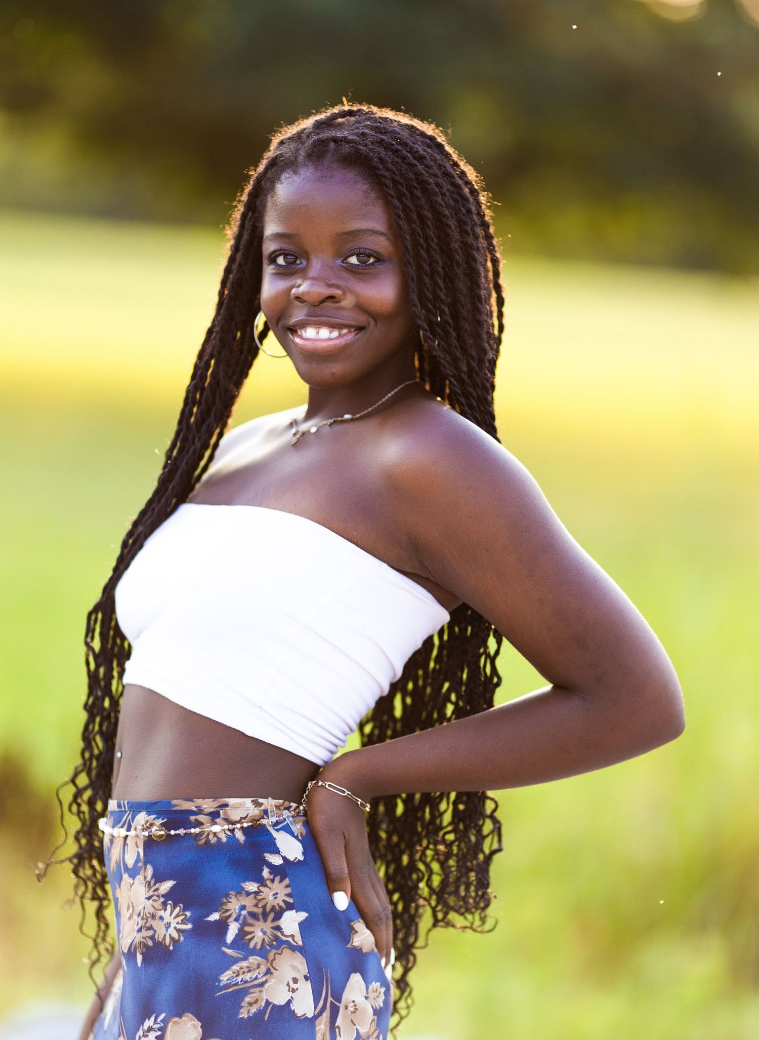 Person with long braids and white crop top poses against blurred nature background in golden sunlight.