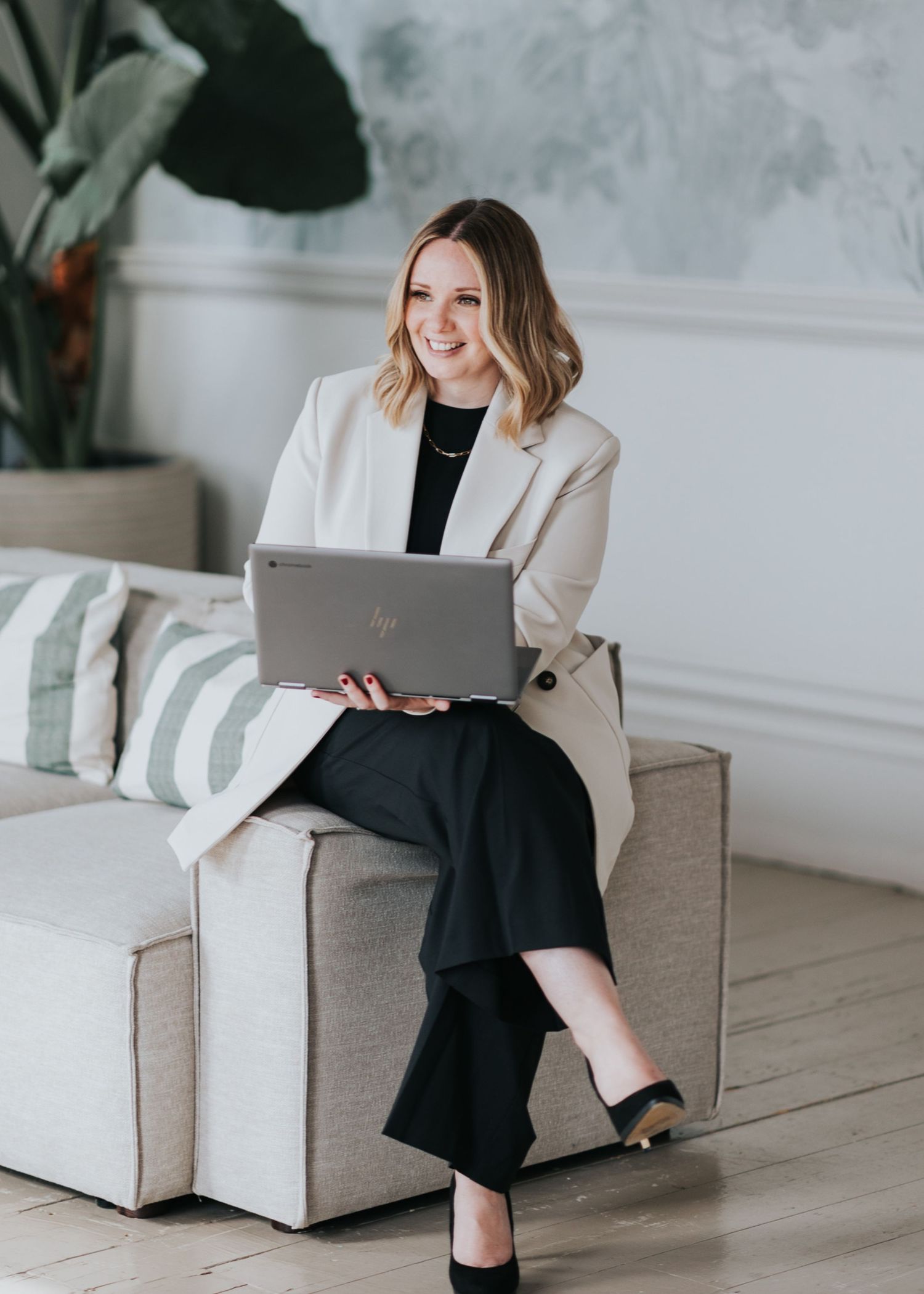 Professional woman in business attire sits with laptop on gray couch in modern minimalist office setting. The Bugle Headshots Southampton