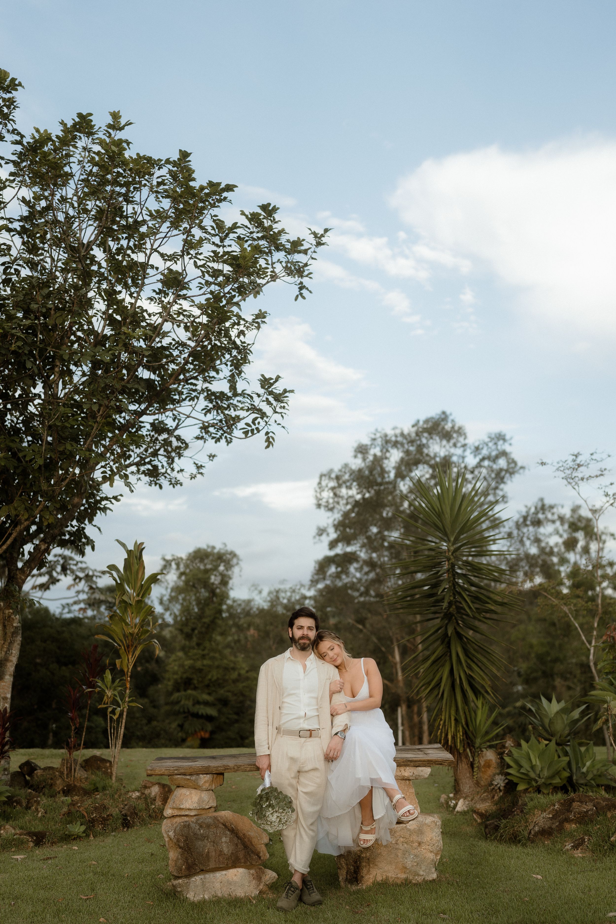 Couple in wedding attire posing on stone wall in tropical garden setting.
