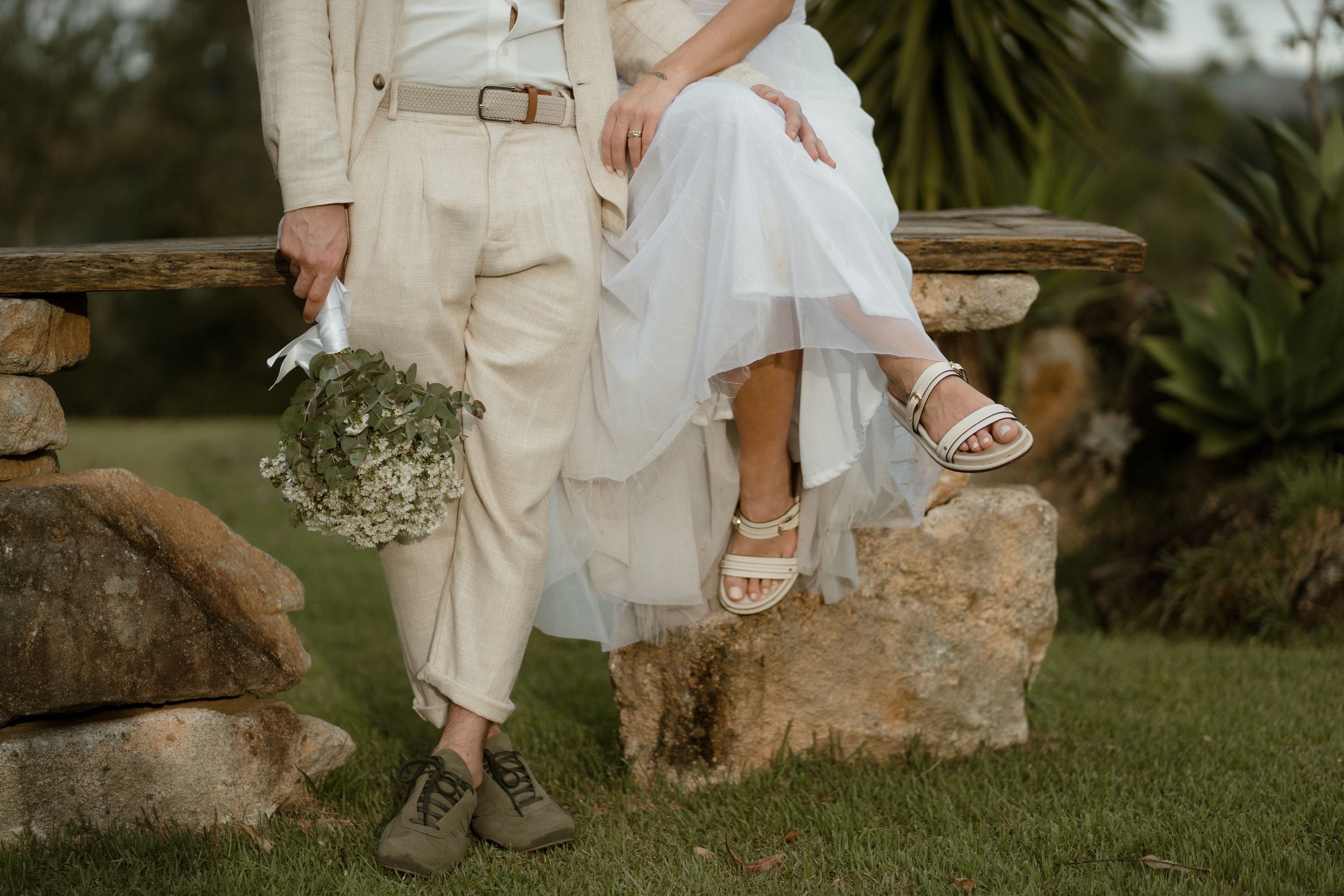 Detail shot of couple's legs sitting on stone wall with wedding dress and bouquet.