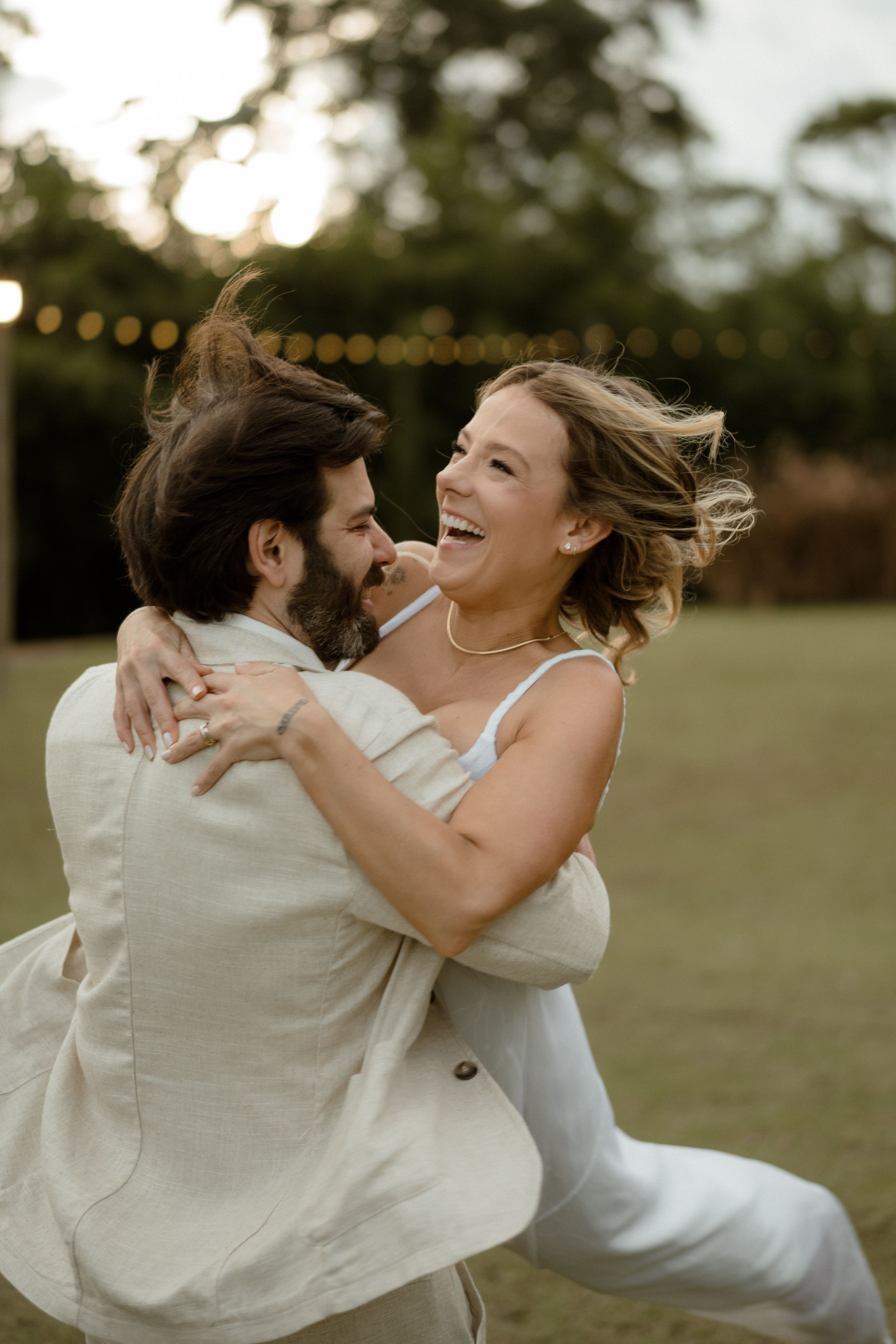 A couple dances and laughs together outdoors at sunset with string lights in the background.