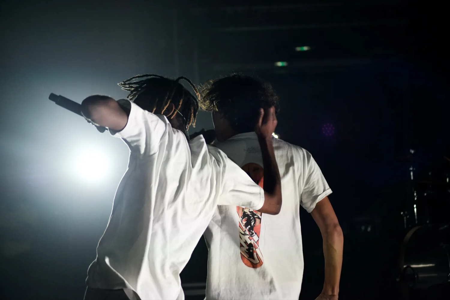 Deux rappeurs sur scène, de dos, bras levés sous un projecteur intense, ambiance de concert énergique.