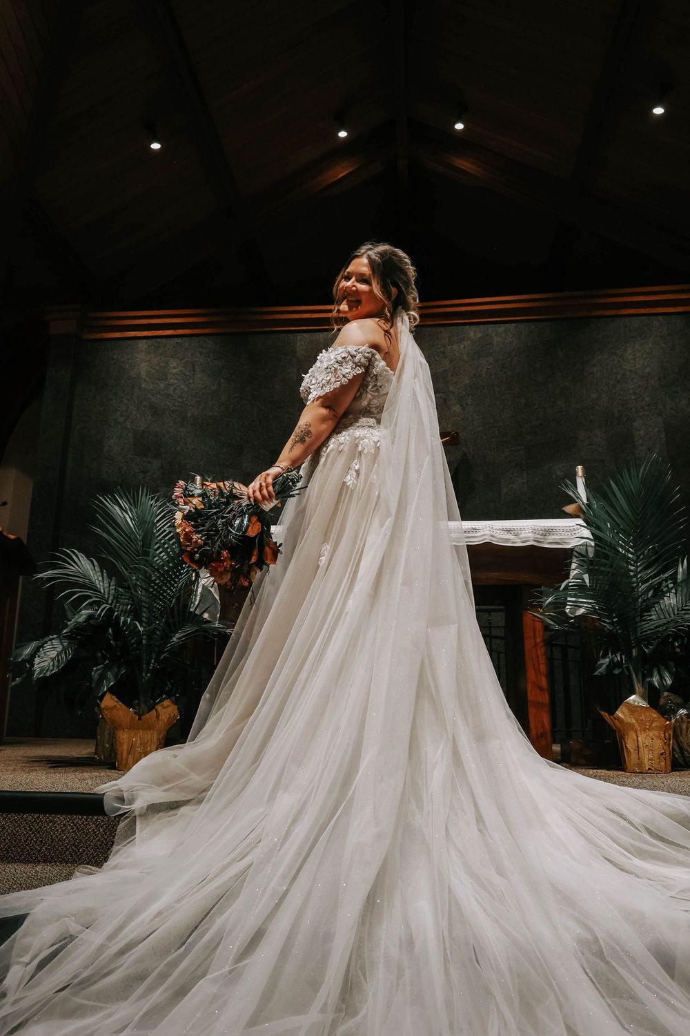 A bride in a flowing white gown with a dramatic long train poses while holding a bouquet in a dimly lit venue with palm plants.