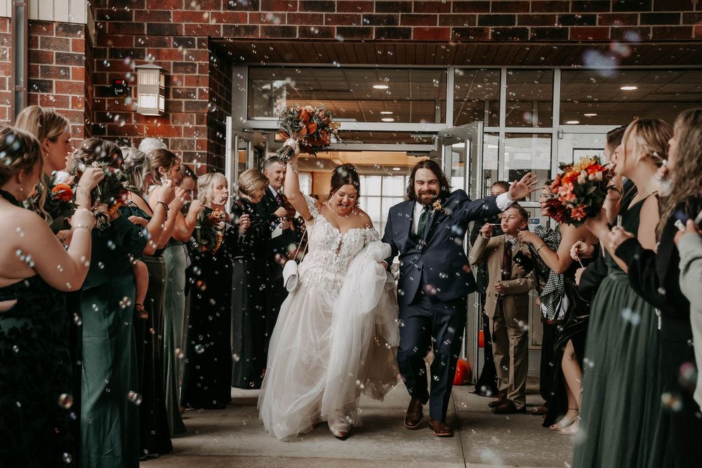 Wedding guests in emerald green dresses throw confetti as couple walks through rustic brick venue corridor.