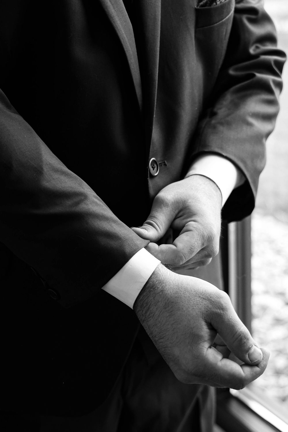 Close-up black and white shot of groom adjusting his suit cuff by a window.