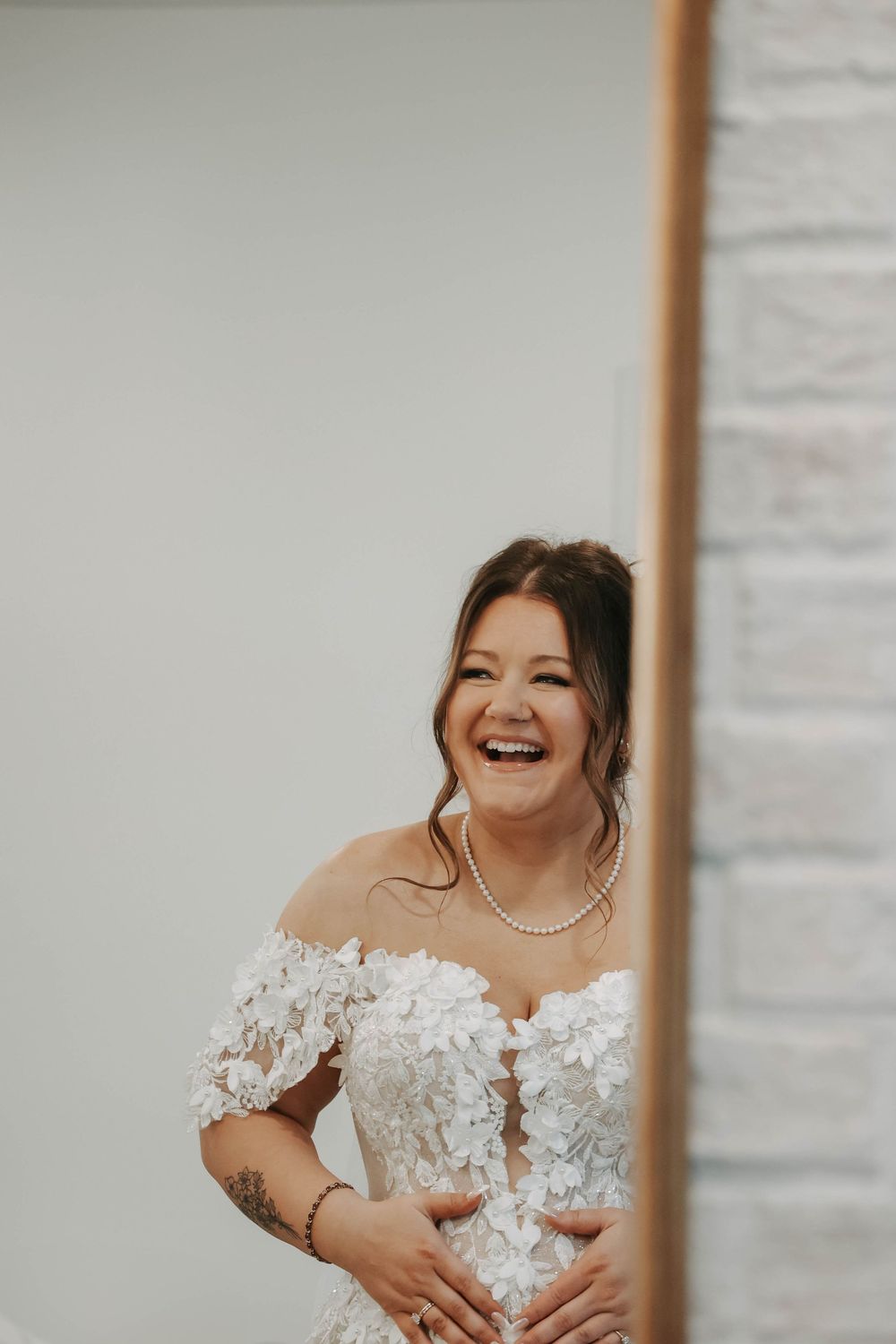 A joyful bride wearing an off-shoulder lace wedding gown with floral appliques and a pearl necklace.