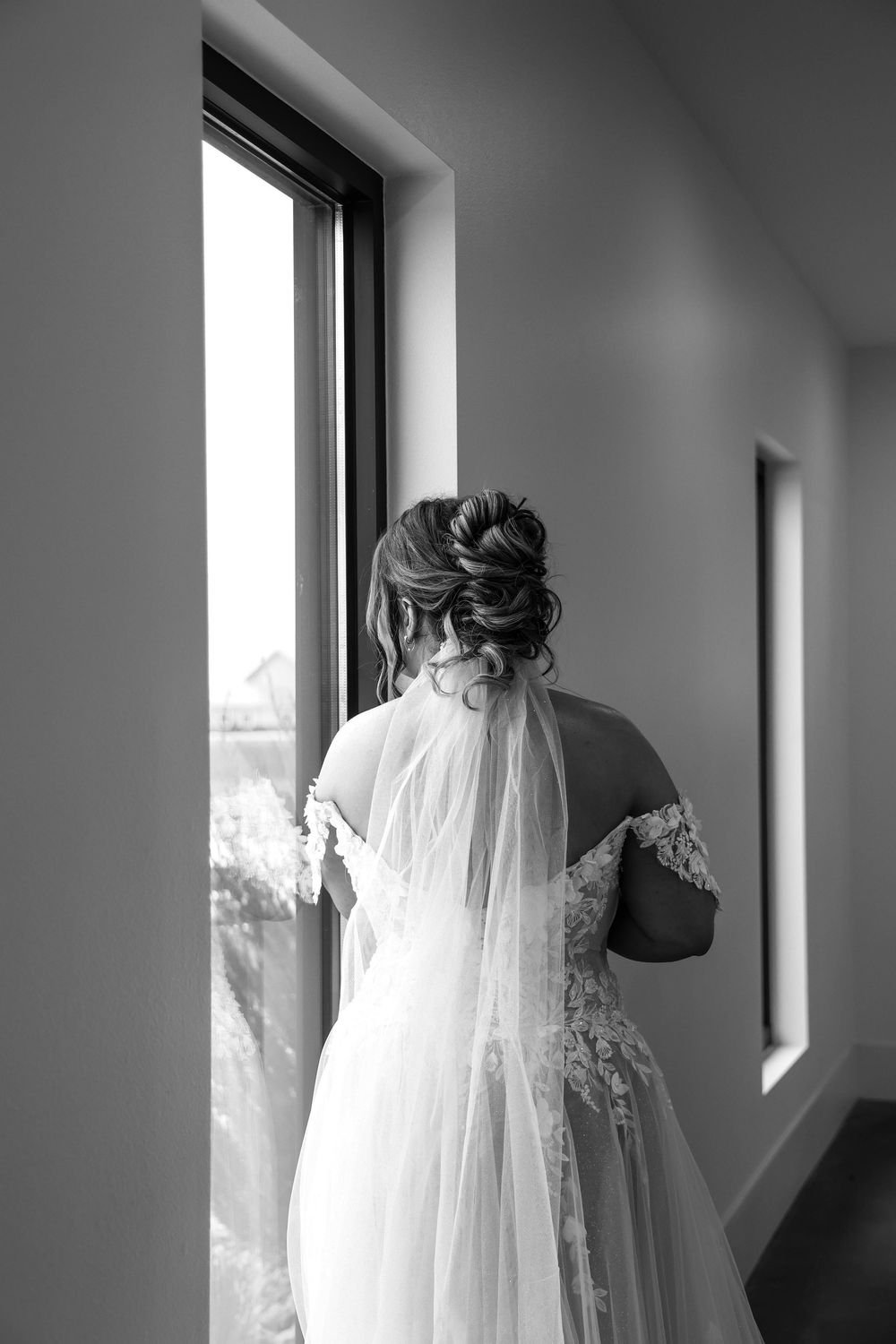 Black and white photo of a bride in a lace wedding dress and veil looking out a window.