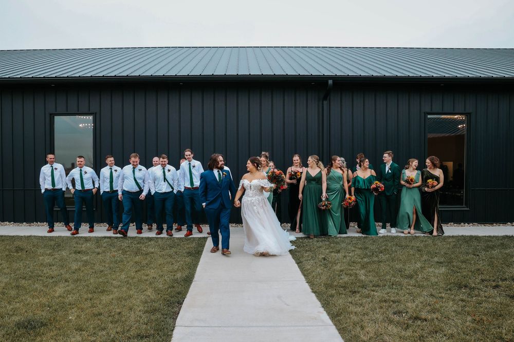 Wedding party poses on white walkway with bridesmaids in green dresses and groomsmen in light blue shirts.