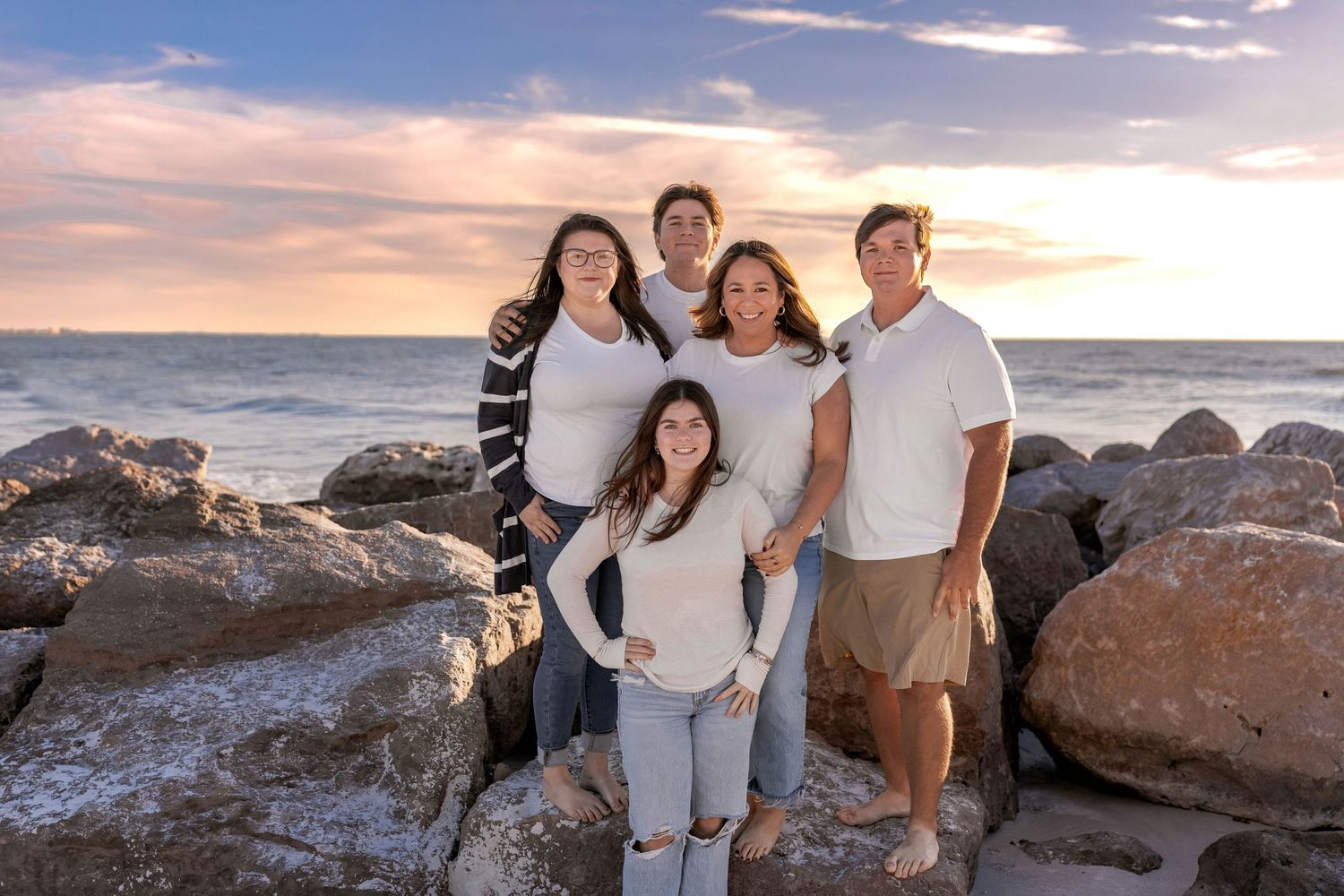 Family portrait session on rocky beach during golden hour with ocean waves and sunset.