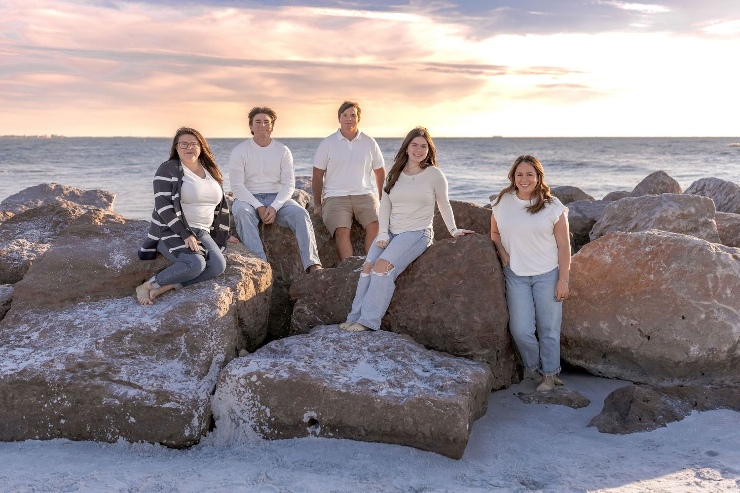 Family sitting on coastal rocks during sunset with dramatic pink and orange sky.