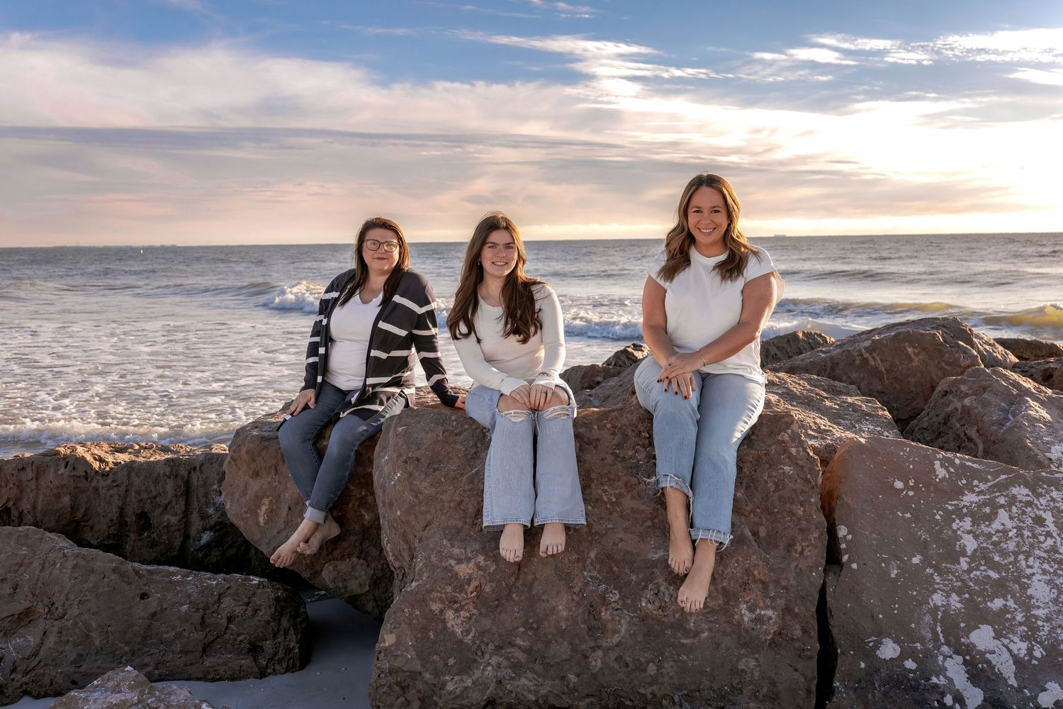 Family members posed on coastal rocks with dramatic sunset sky and ocean waves behind them.