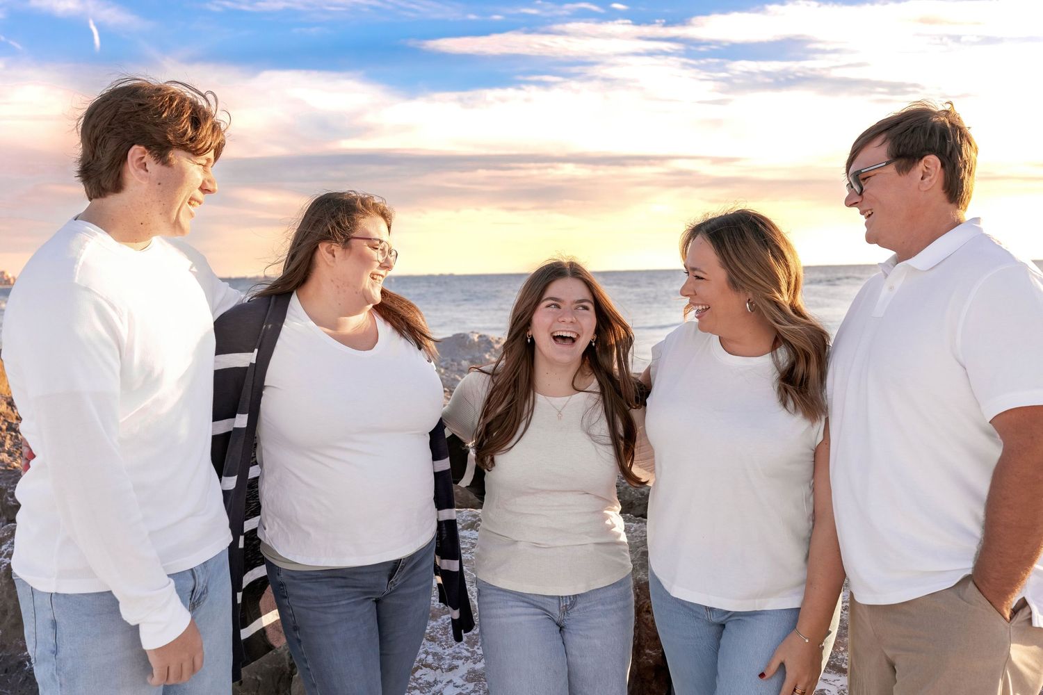 Five people wearing matching white outfits share a candid moment of laughter at sunset on a beach.
