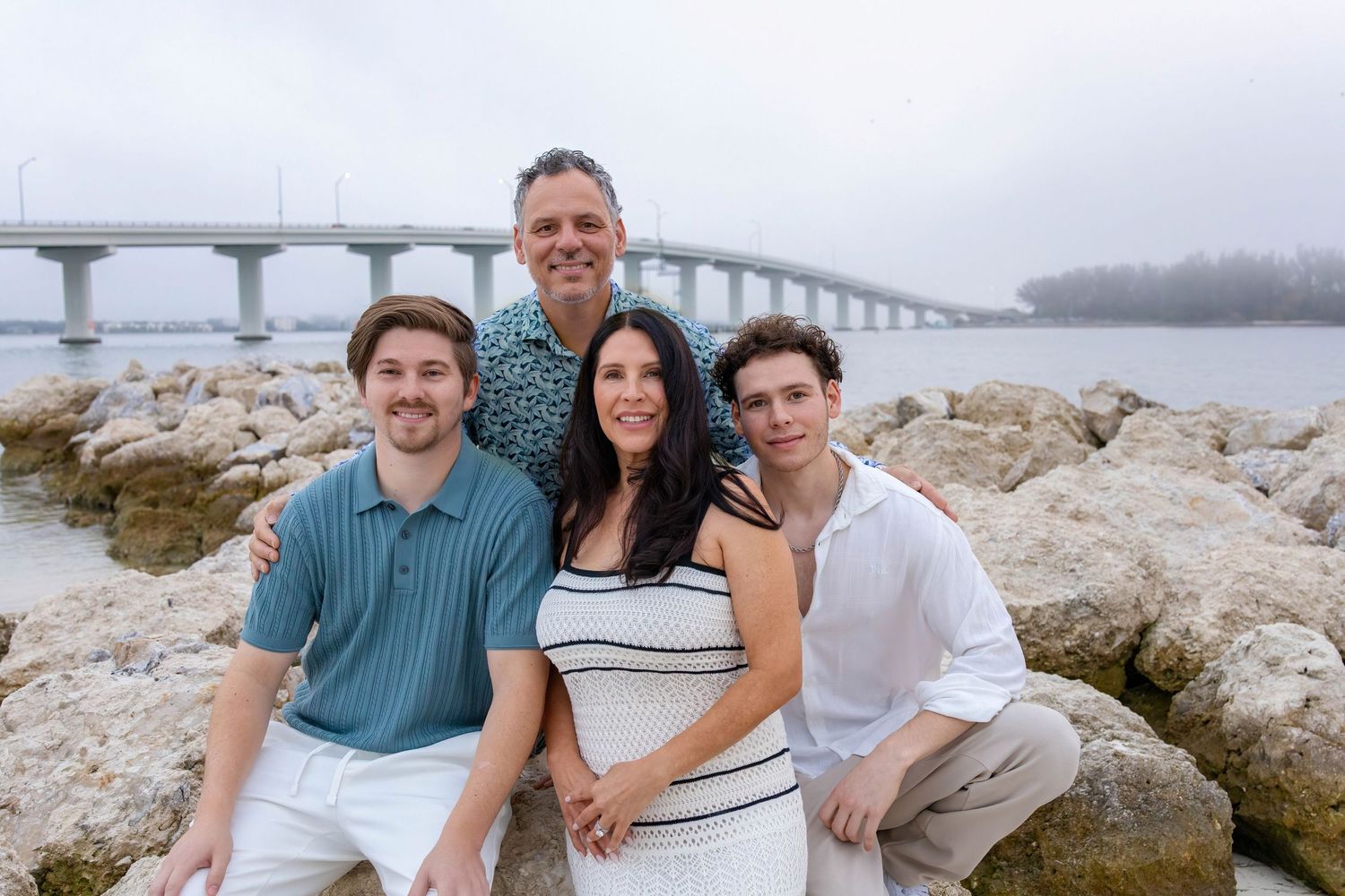 Family group photo on rocky beach with bridge visible in background under cloudy skies.