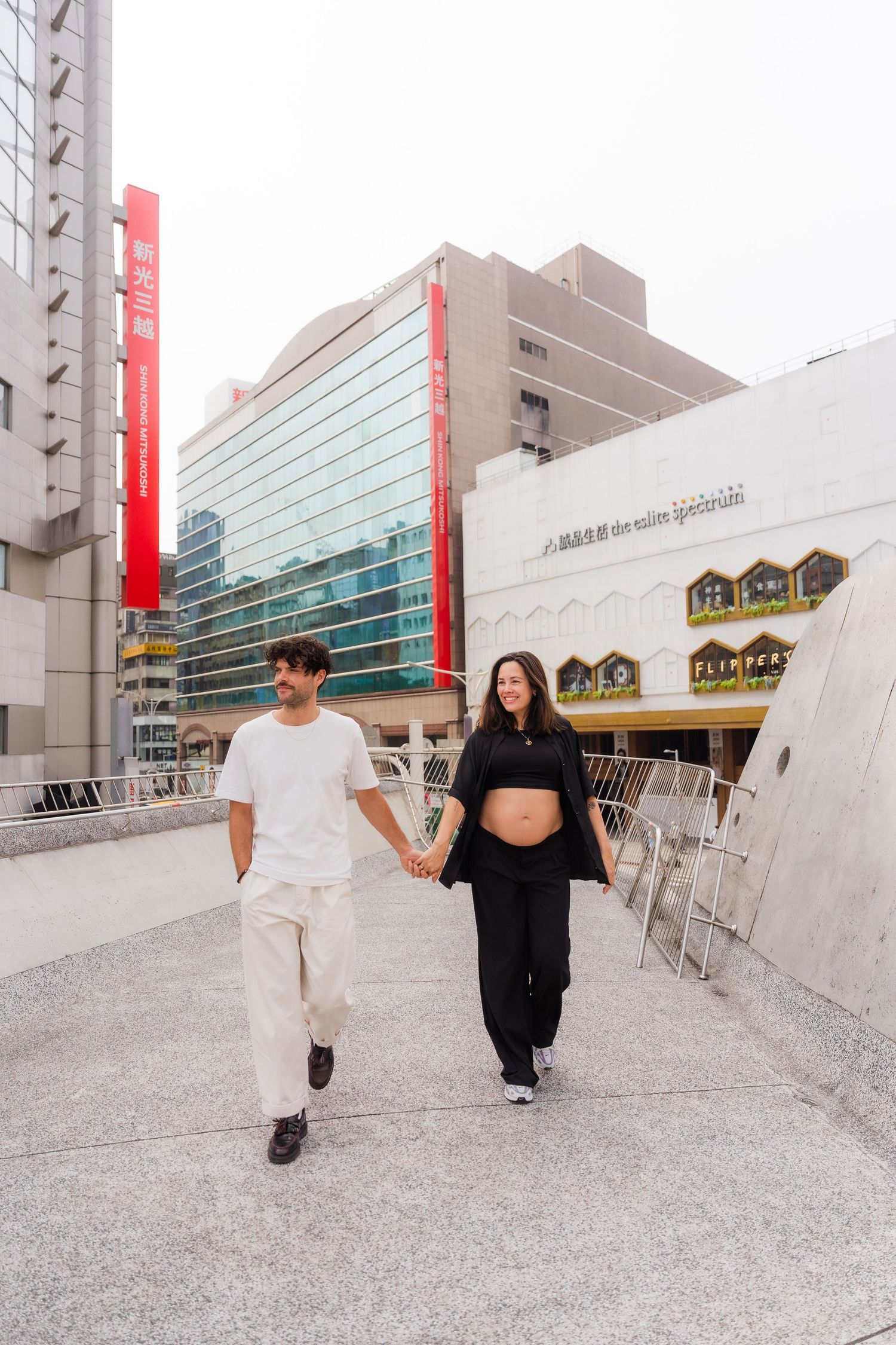 A couple in coordinated white and black outfits walk hand in hand outside a modern building with red accents.