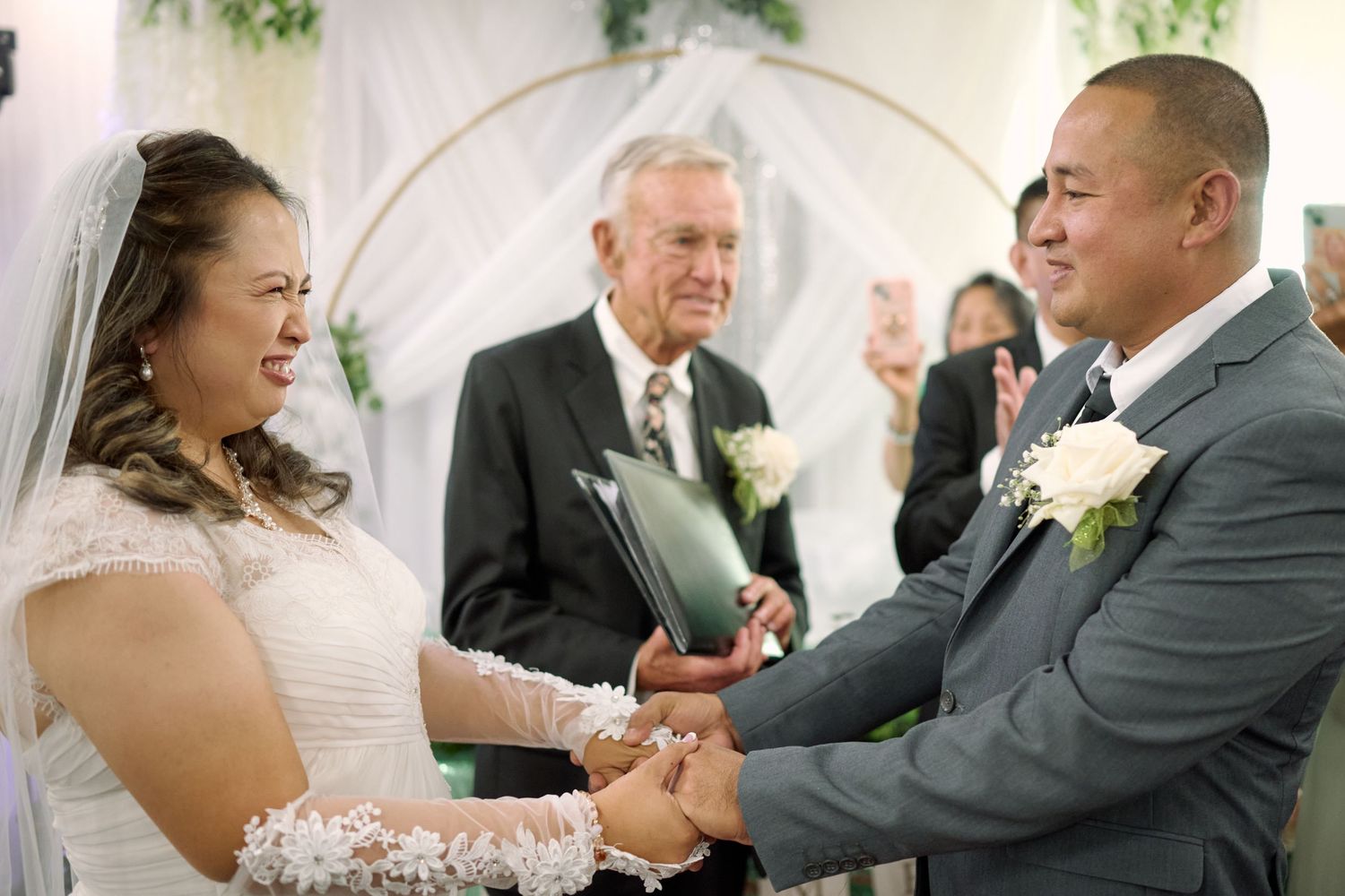 A joyful wedding ceremony takes place under a white arch with two people holding hands and an officiant in the middle.