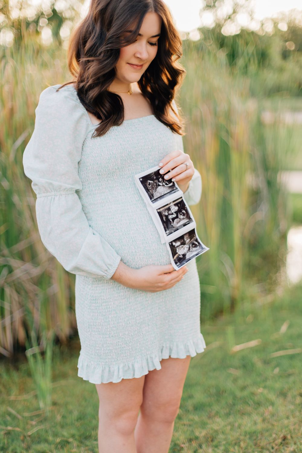 A pregnant woman in a white dress holds ultrasound photos while standing in a grassy field.