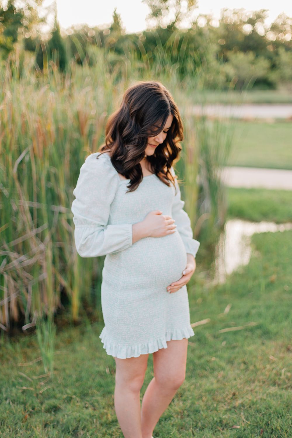 A pregnant woman in a white ruffled dress stands in a serene outdoor setting with tall grass and natural lighting.