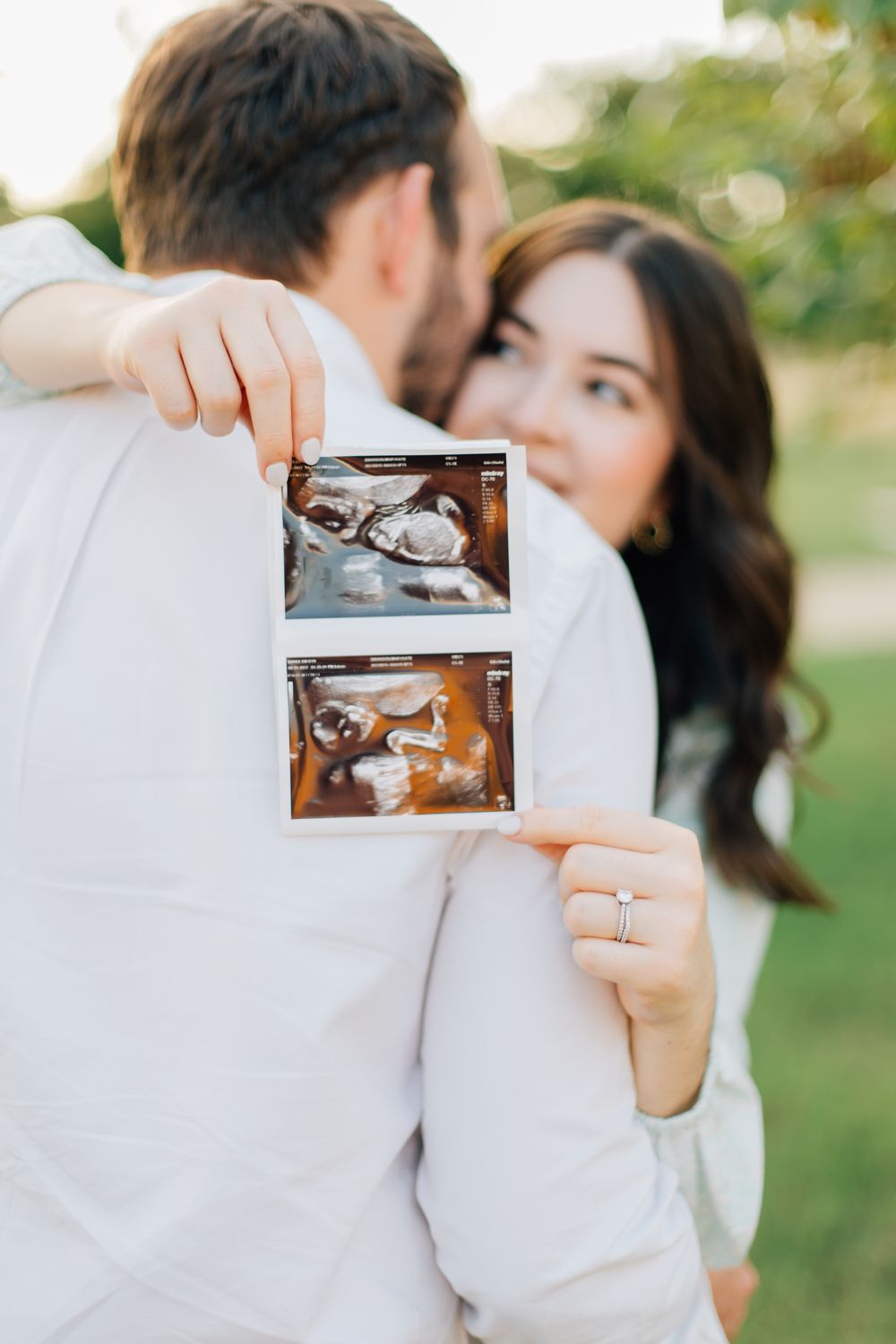 A couple holds up ultrasound images while embracing outdoors in a sweet pregnancy announcement moment.